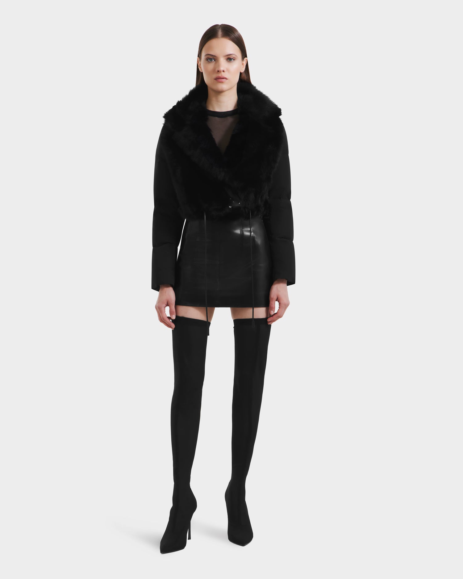 A woman stands facing forward against a plain light background, wearing the Rudsak LILAH - 8125509 Black cropped jacket with a black faux fur collar, paired with a black mini skirt, sheer tights, and black over-the-knee heeled boots.