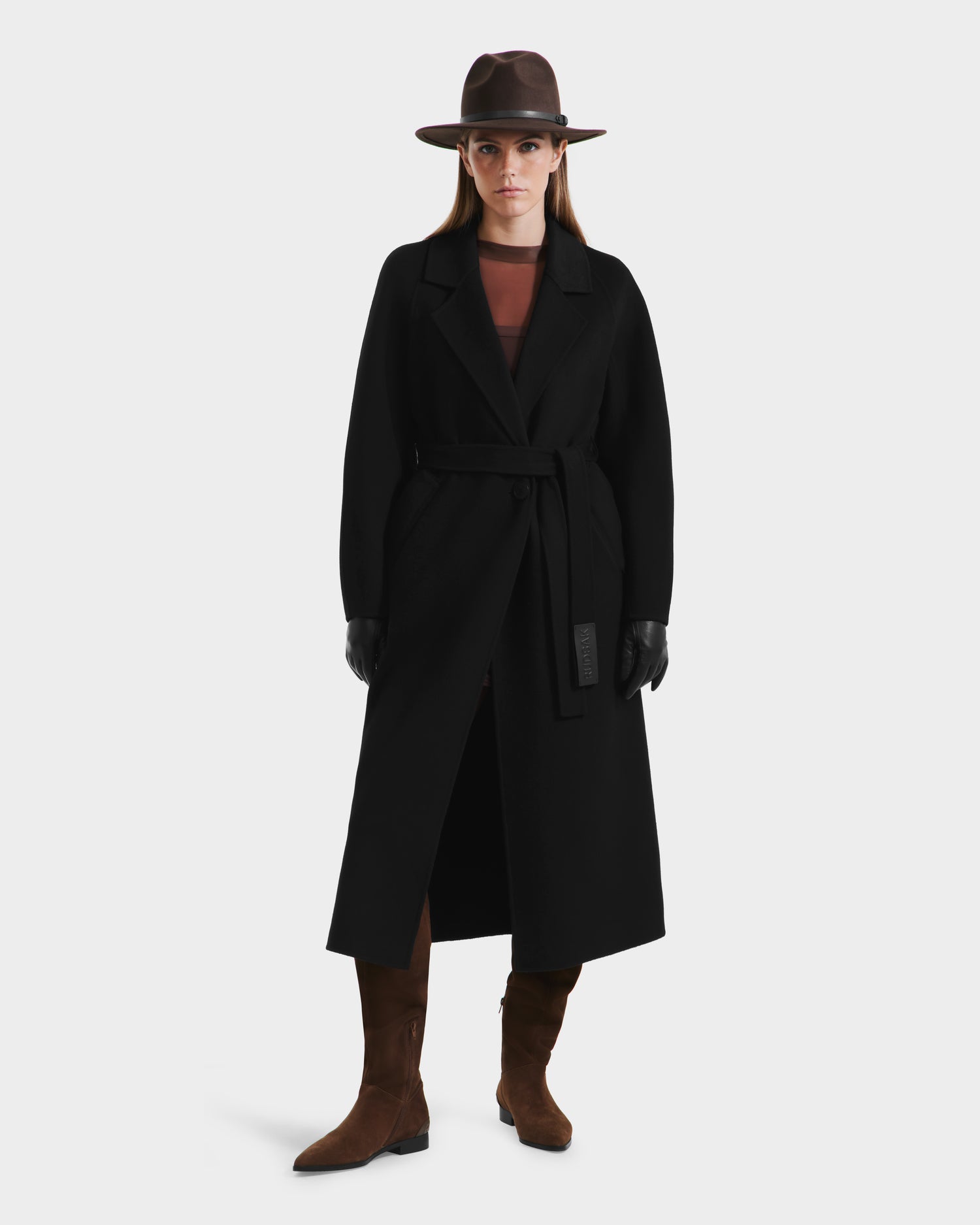 A person is wearing the Rudsak LILIA - 8125634 Black womens long wrap coat, styled with a brown hat, boots, black gloves, and a sheer brown top, standing facing forward against a plain white background.