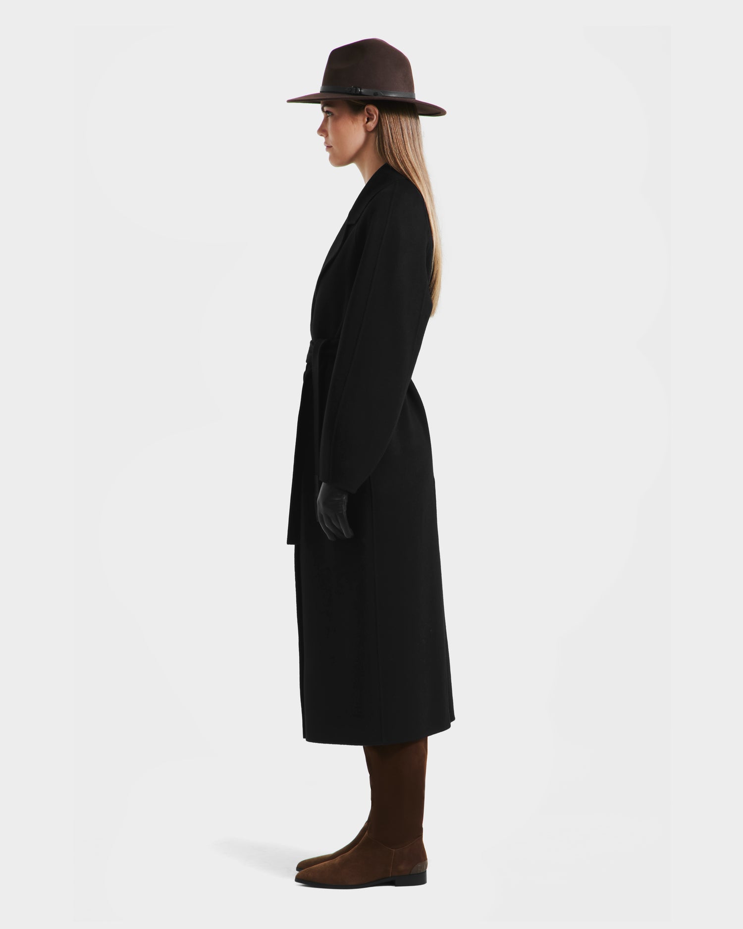 A woman stands in profile against a white background, wearing the Rudsak LILIA - 8125634 Black womens long wrap coat, paired with a dark wide-brimmed hat, gloves, brown knee-high boots, and her long hair down. The perfect transitional layering piece.