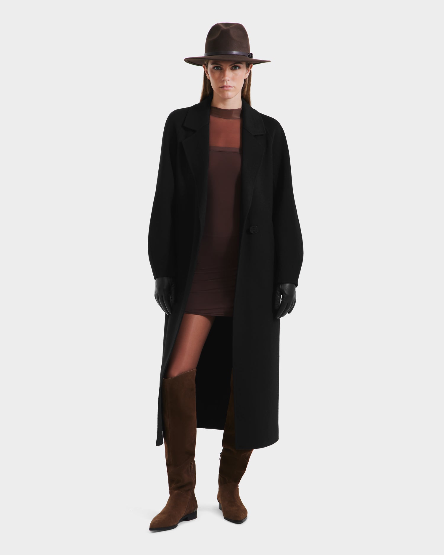 A woman in a Rudsak LILIA - 8125634 Black long wrap coat over a sheer brown dress, with a brown hat, black gloves, and tall brown suede boots stands against a plain white background.