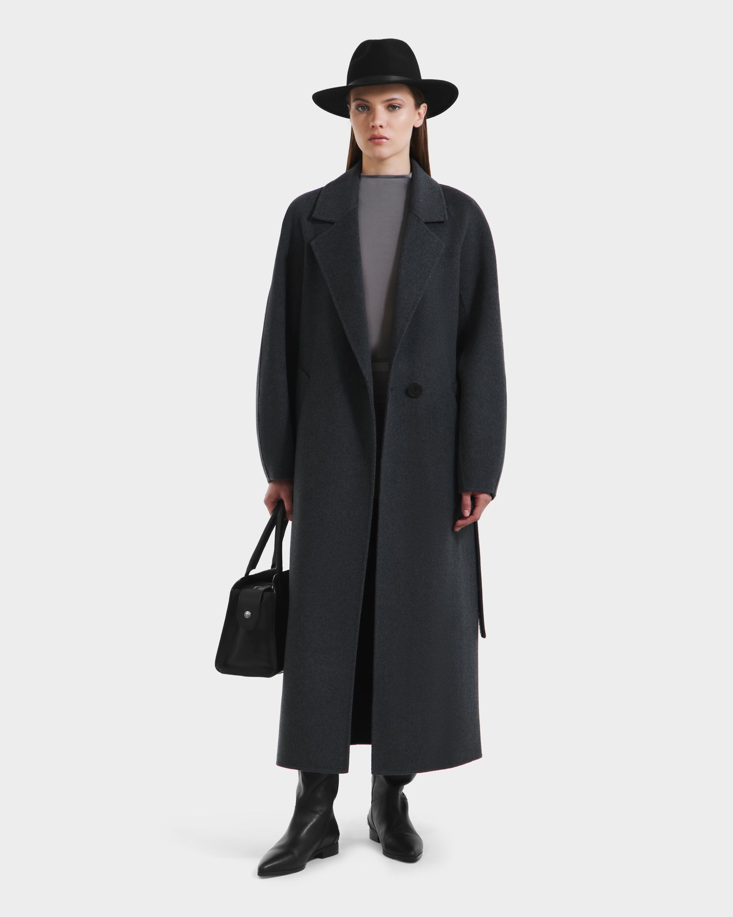 A woman wearing the Rudsak LILIA - 8125634 Heather Charcoal long wrap coat over a gray top, with a wide-brim black hat, black boots, and a black handbag stands facing forward against a plain white background.