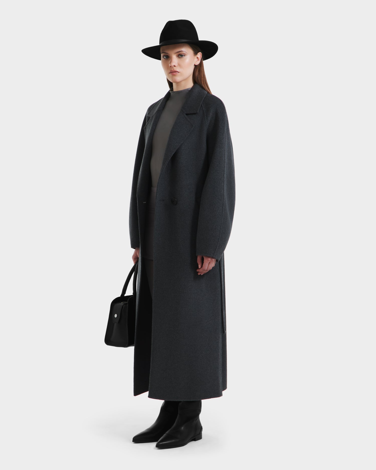 A woman wears the Rudsak LILIA - 8125634 Heather Charcoal coat with a black wide-brim hat, gray turtleneck, black ankle boots, and holds a black handbag against a plain light background.