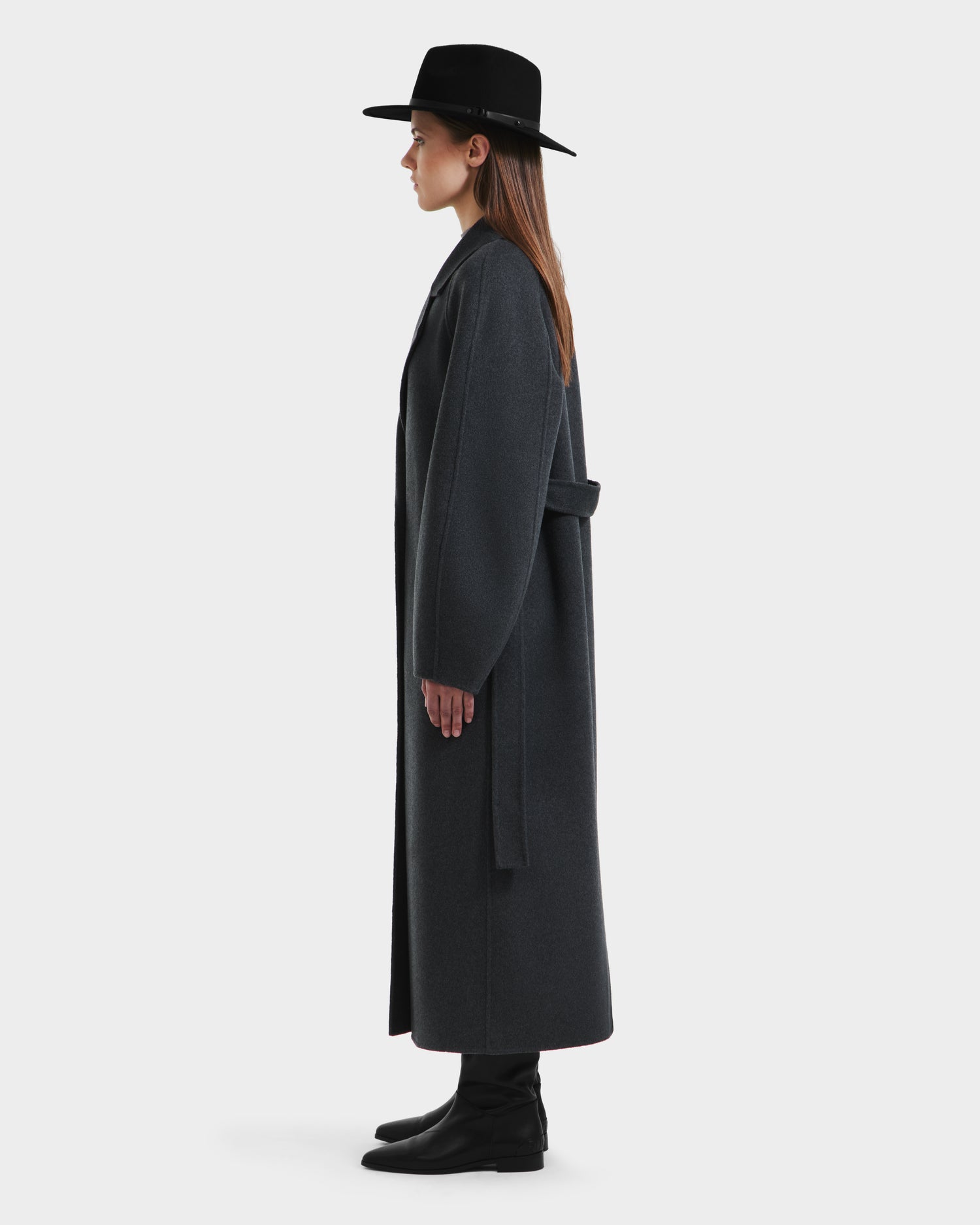 A woman stands in profile against a plain light background, wearing the Rudsak LILIA - 8125634 Heather Charcoal long wrap coat, black boots, and a black wide-brimmed hat.