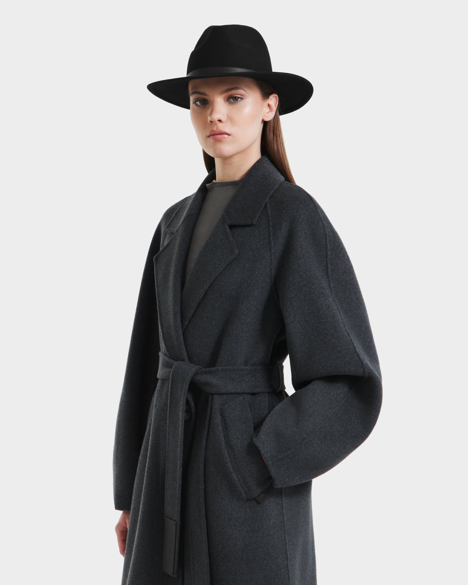 A woman in a dark wide-brim hat and the Rudsak LILIA - 8125634 Heather Charcoal belted women’s long wrap coat stands against a white background, looking at the camera with a neutral expression.