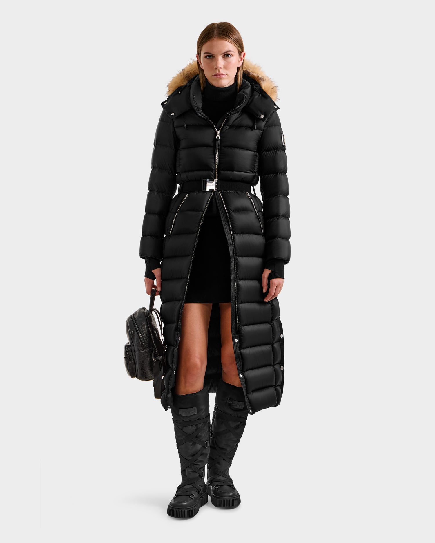 A woman in the Rudsak LILITH L - L125528 Black maxi-length down puffer with a fur-trimmed hood, black turtleneck, short skirt, knee-high boots, and holding a black backpack stands facing forward against a plain background.
