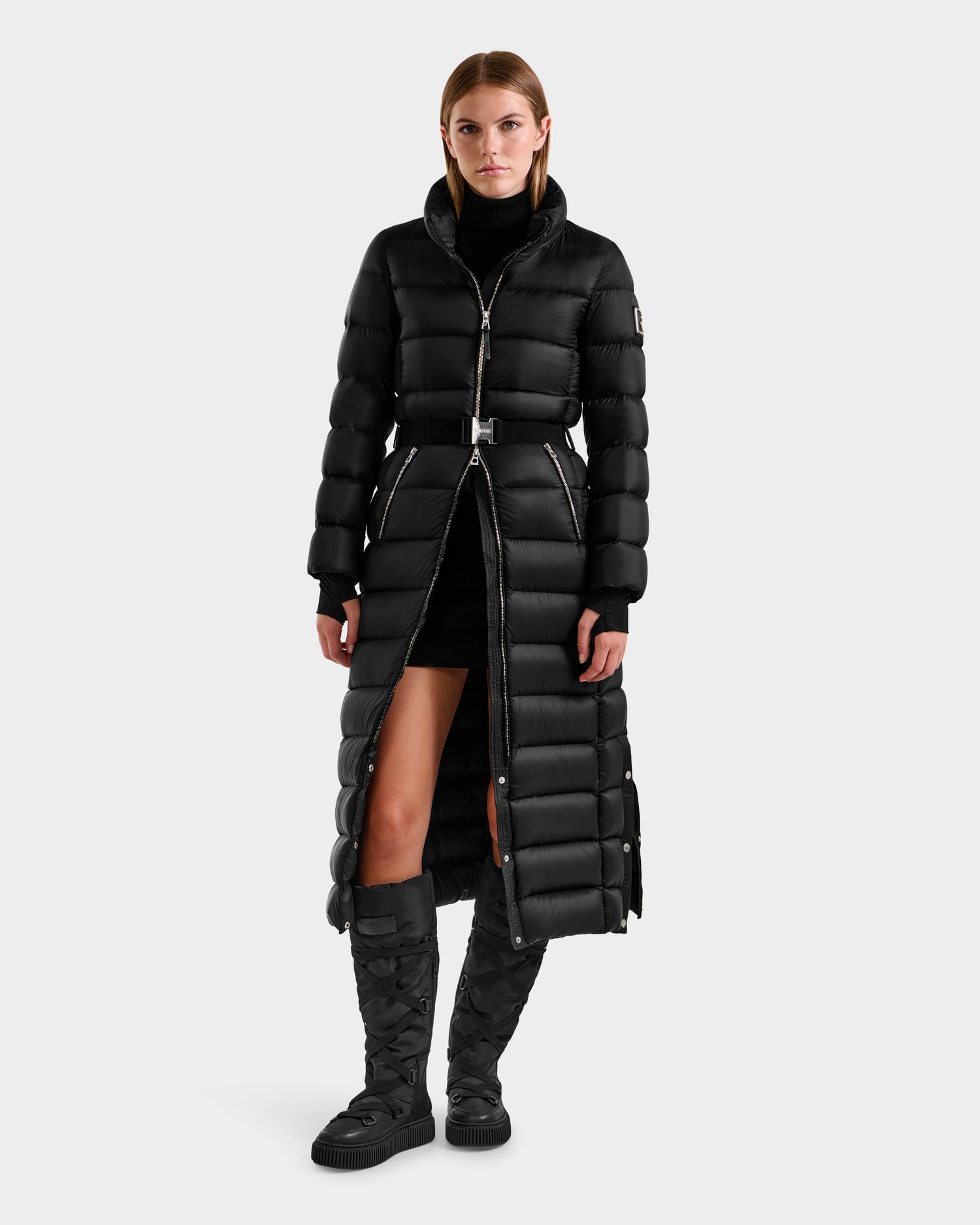 A woman faces forward in a Rudsak LILITH L - L125528 Black maxi-length down puffer with RDS-certified filling and removable belt, styled with a black turtleneck, short skirt, and quilted knee-high boots against a plain white background.