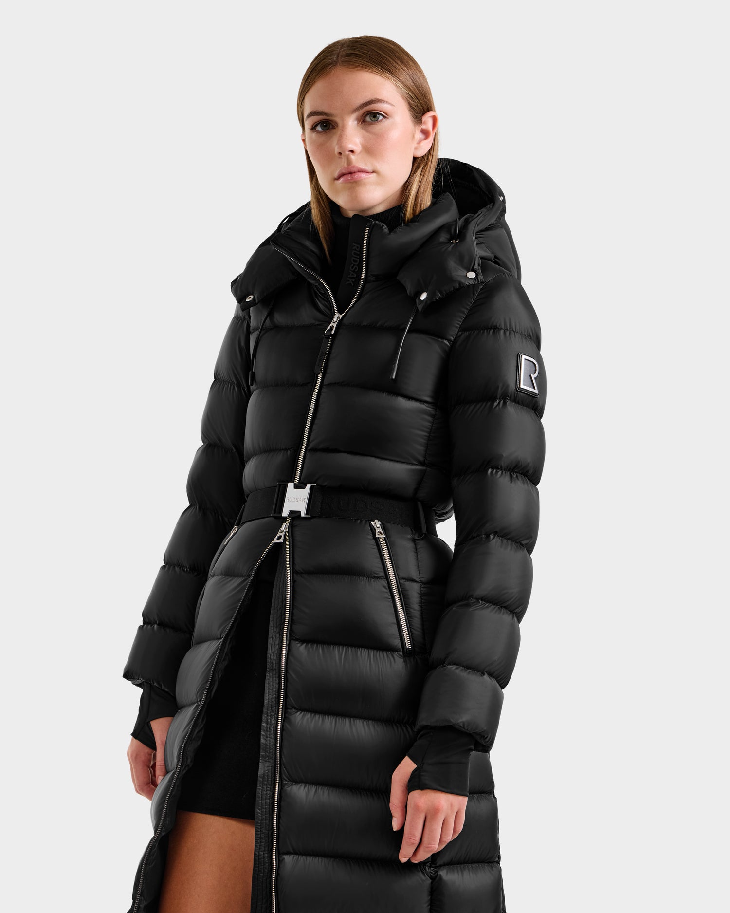 A woman faces forward wearing the Rudsak LILITH L - L125528 Black maxi-length down puffer jacket with hood, removable belt, zippered pockets, and an "R" patch on the upper sleeve.