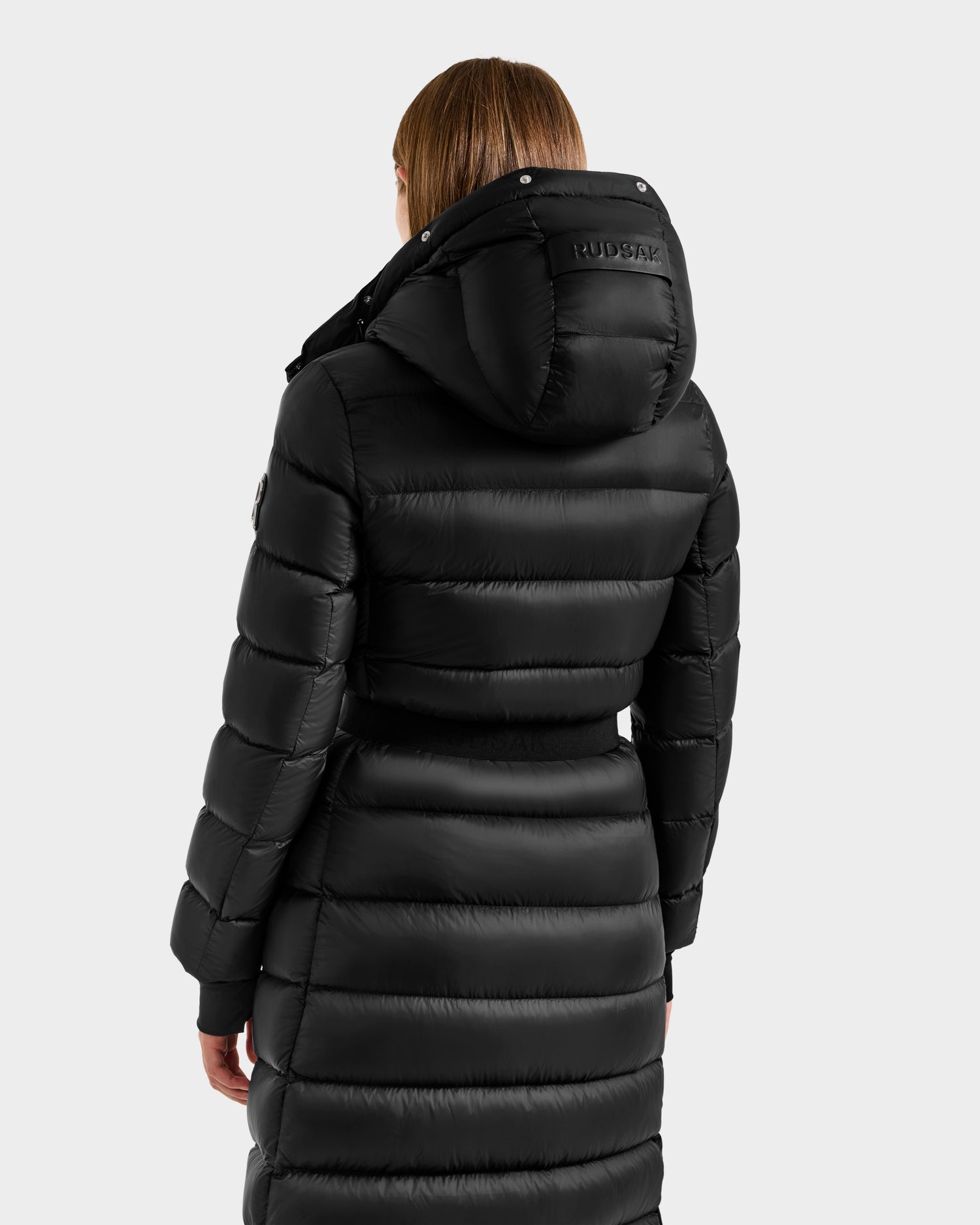 A person wearing the Rudsak LILITH L - L125528 Black maxi-length down puffer coat with a hood, horizontal quilting, and RUDSAK branding on the hood, viewed from the back against a plain light gray background. Filled with RDS-certified down.