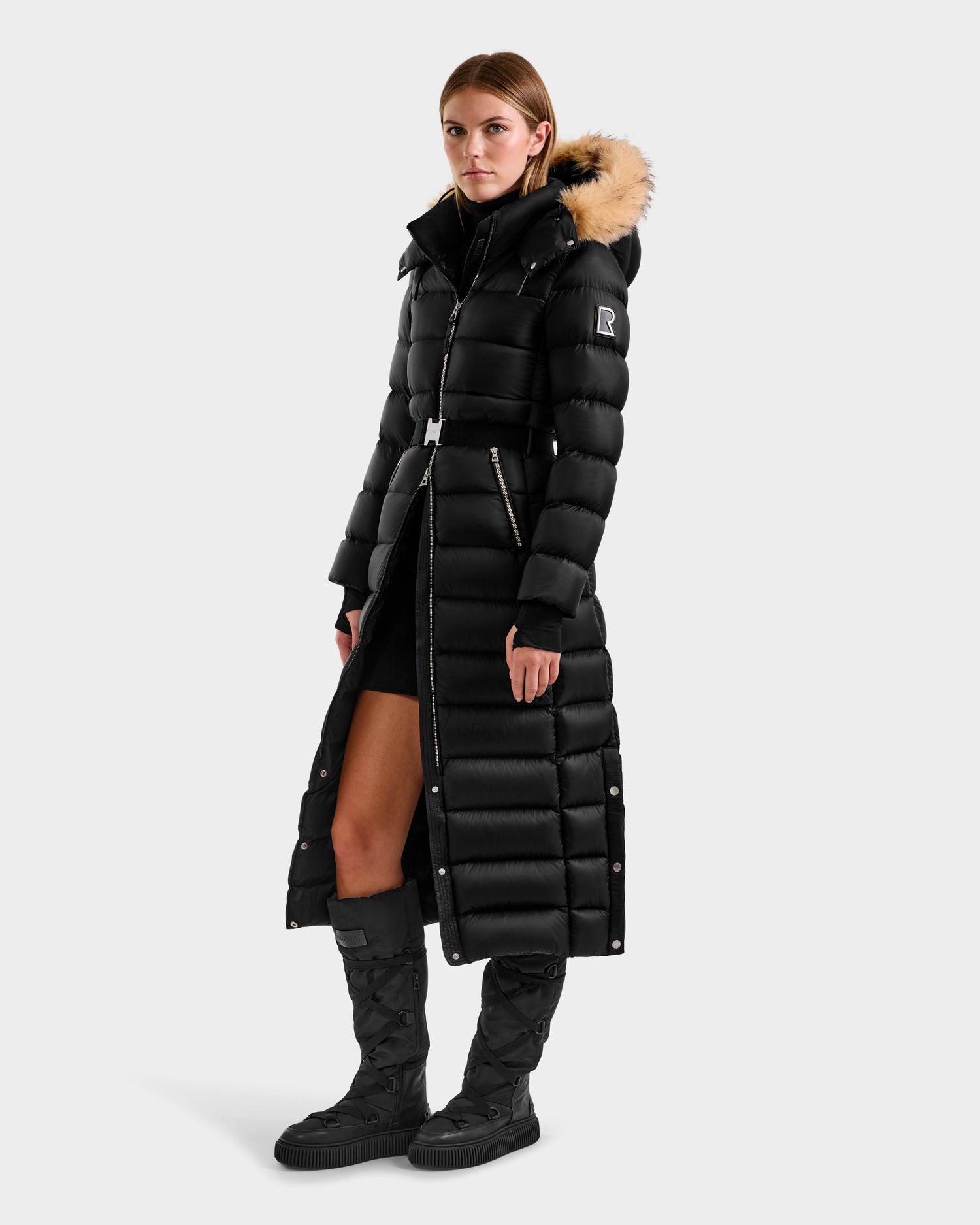 A woman models the Rudsak LILITH L - L125528 Black maxi-length down puffer coat with a fur-trimmed hood and removable belt, paired with black knee-high quilted boots and a black outfit, standing slightly sideways against a light background.