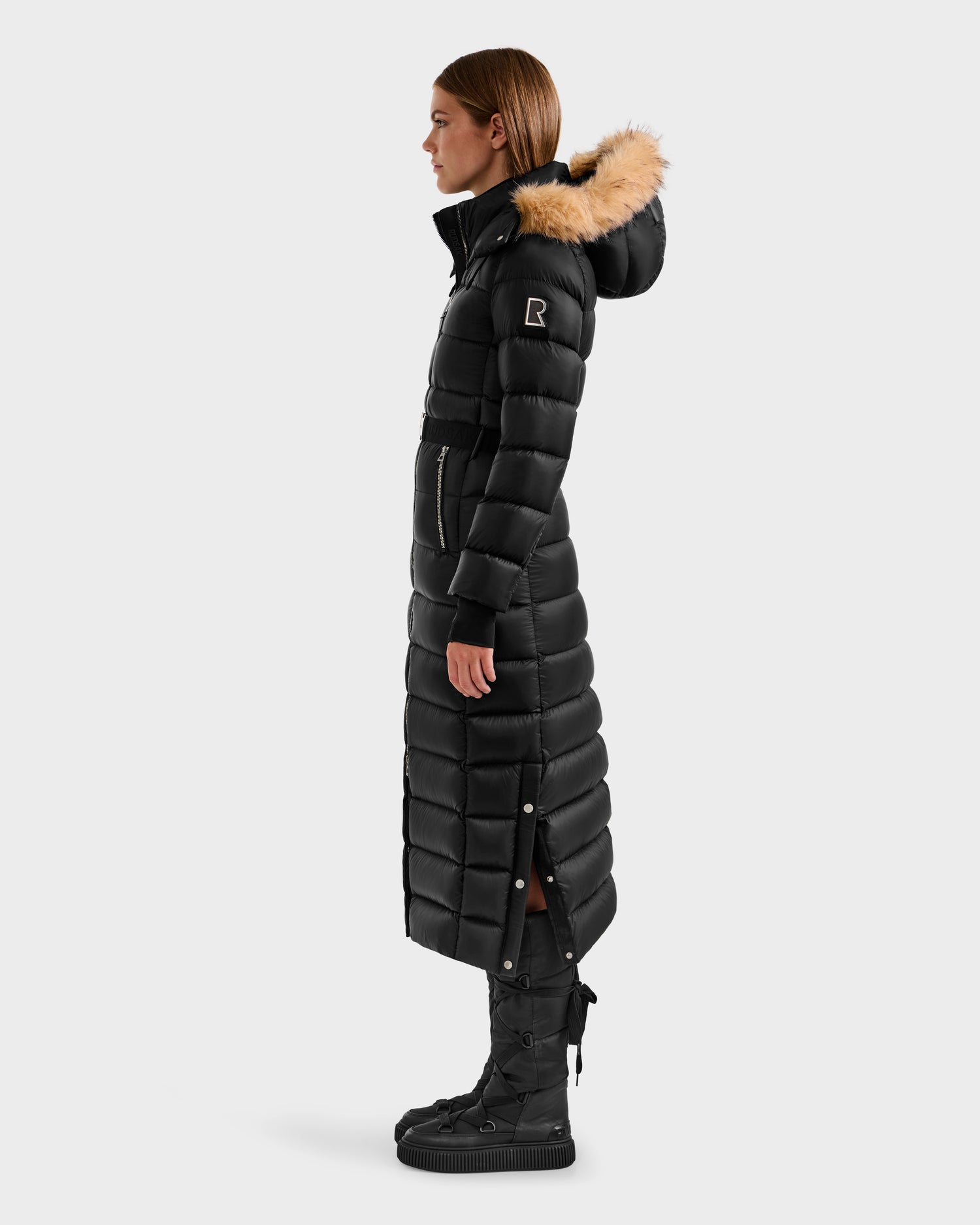 A person stands in profile wearing the Rudsak LILITH L - L125528 Black maxi-length down puffer with a fur-trimmed hood and black boots, hands by their sides against a white background. The coat features RDS-certified down for warmth and ethical comfort.