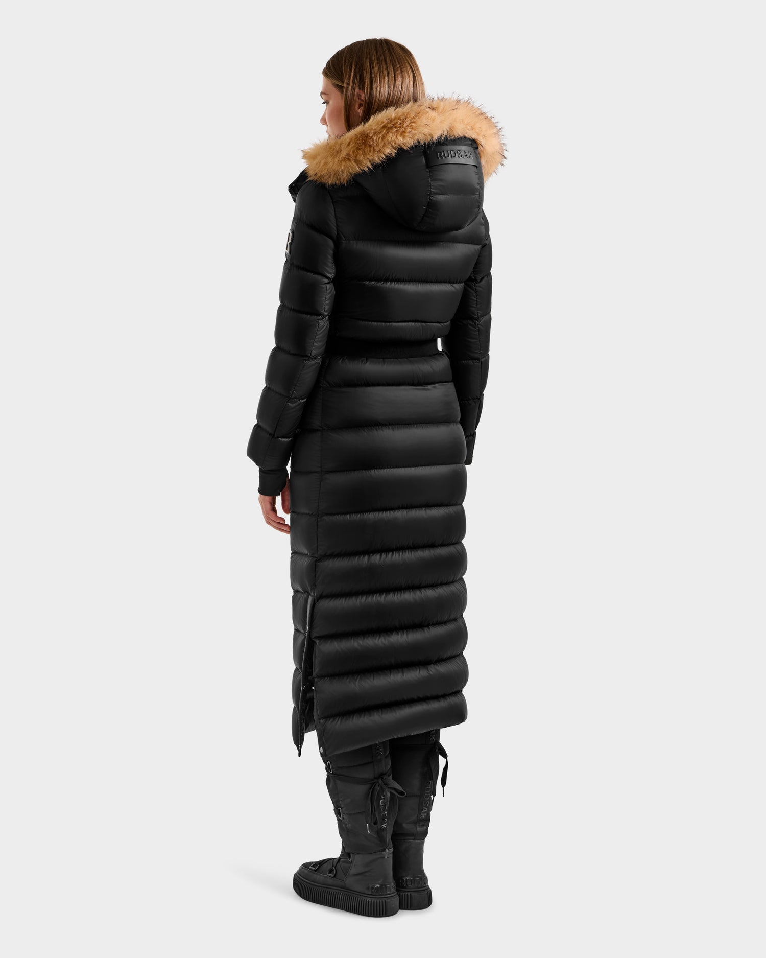 A woman faces away, wearing the Rudsak LILITH L - L125528 Black maxi-length down puffer with a fur-trimmed hood, black boots, and black pants. The RDS-certified coat extends to her calves and has horizontal quilting.