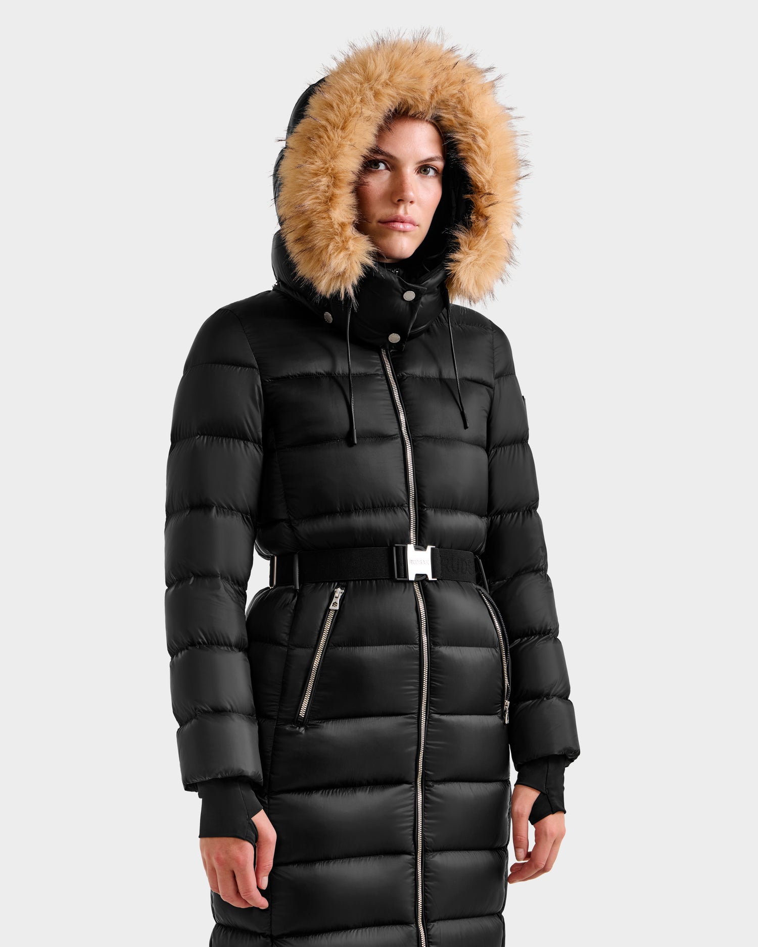 A woman wears the Rudsak LILITH L - L125528 Black maxi down puffer coat with a faux-fur trim hood and belted waist, standing against a plain light gray background.
