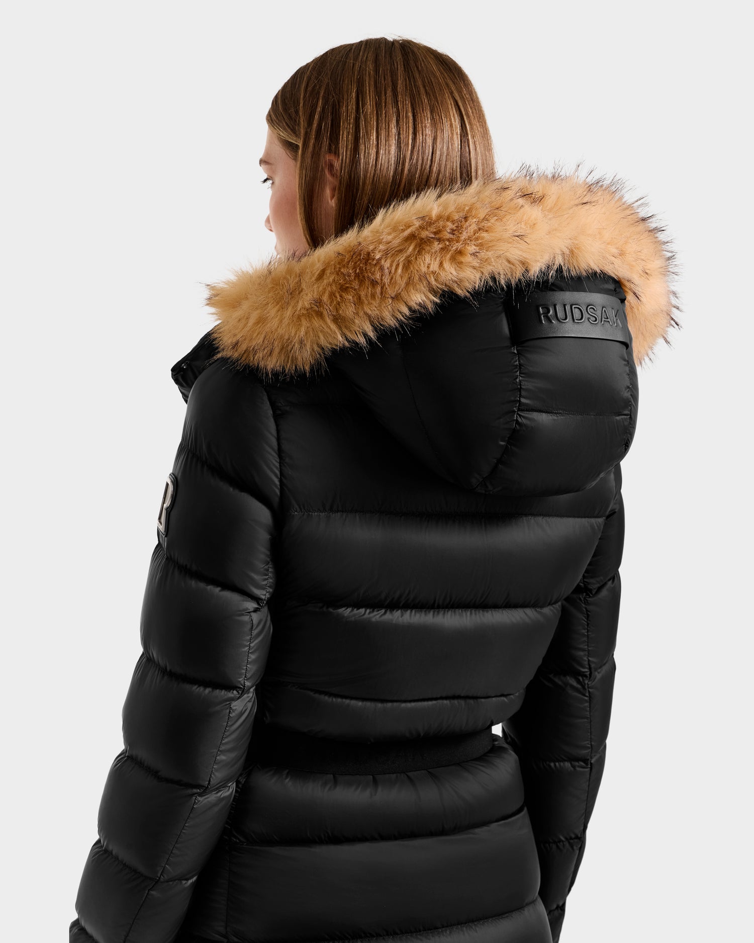 A person with straight, light brown hair wears the Rudsak LILITH L - L125528 Black maxi-length down puffer jacket with a tan faux fur-trimmed hood, RUDSAK on the back of the hood, and RDS-certified down filling for warmth.