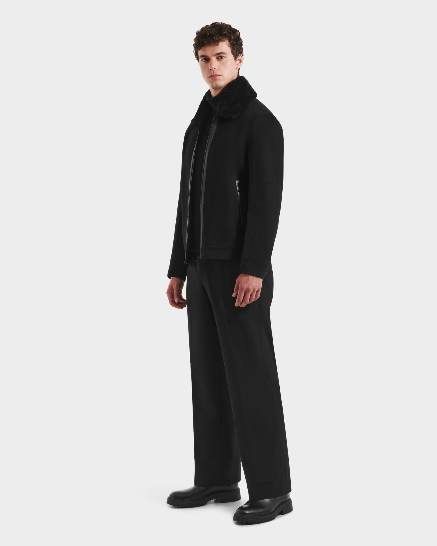 A man with curly hair stands against a light background, wearing the Rudsak LINCOLN - 6125038 Black mens wool coat with a removable faux fur collar, wide-leg black trousers, and black shoes.