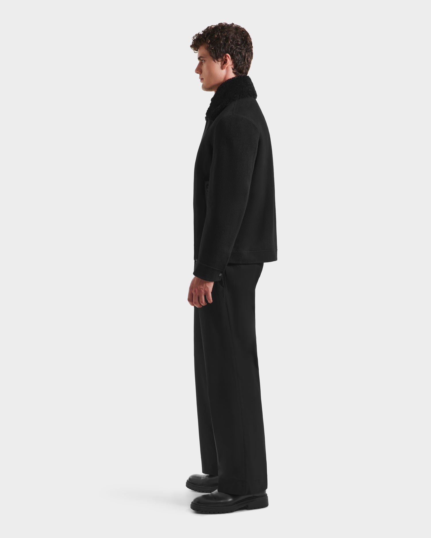 A man with curly hair stands in profile against a white background, wearing the Rudsak LINCOLN - 6125038 Black mens wool coat with a removable faux fur collar, paired with black pants and shoes.