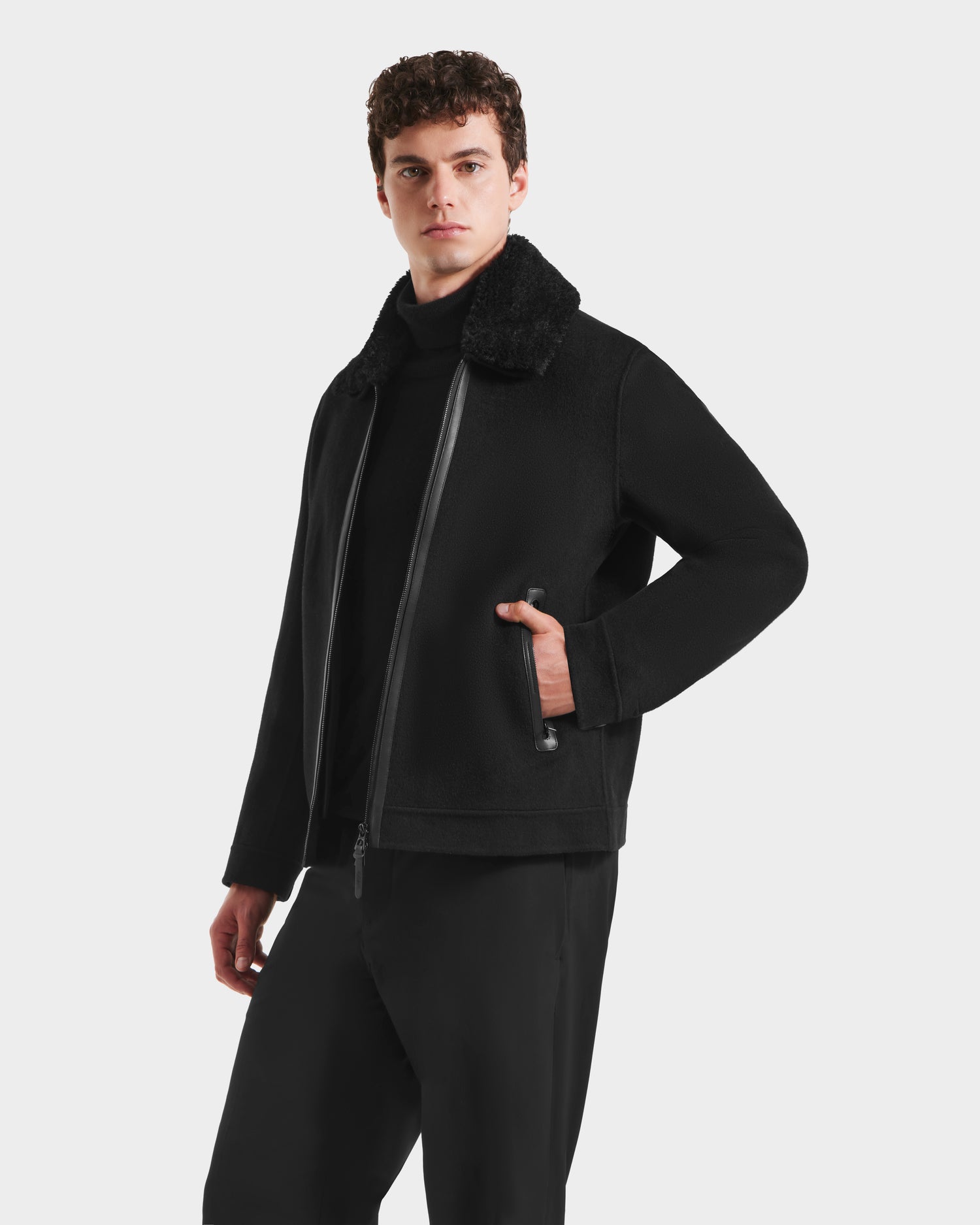 A man wearing the Rudsak LINCOLN - 6125038 Black coat with a removable faux fur collar over a black shirt and pants stands against a white background, looking to the side with one hand in his pocket.