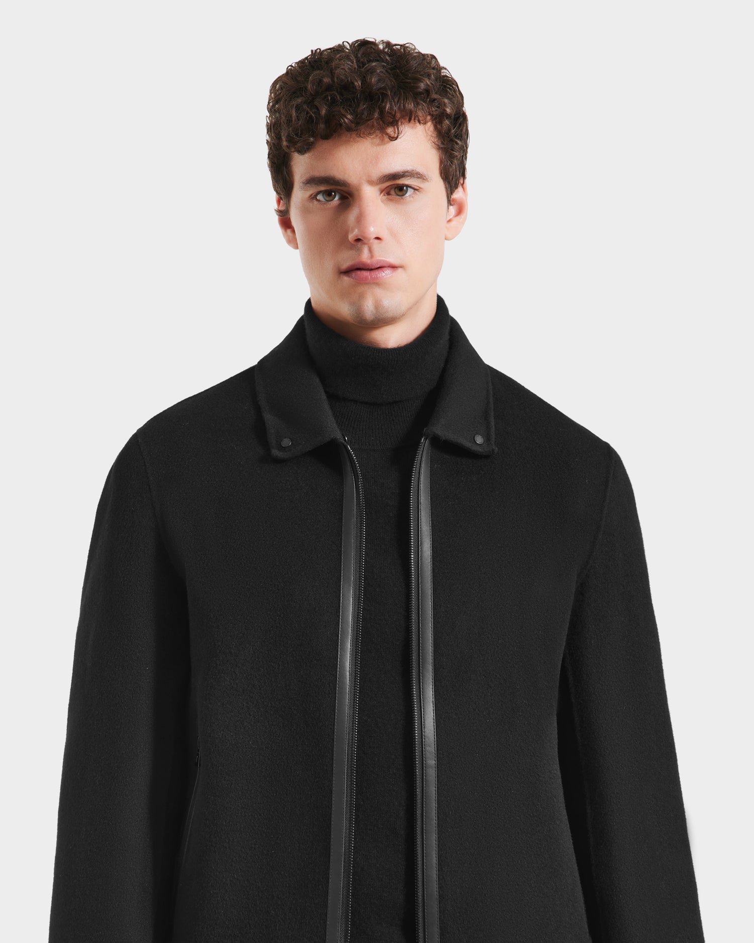 A young man with short curly brown hair wears the Rudsak LINCOLN - 6125038 Black mens wool coat with leather trim and a removable faux fur collar over a high-neck sweater, standing against a plain white background.