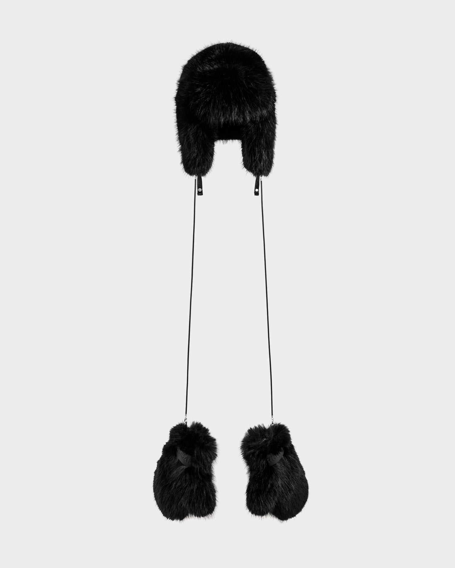 The Rudsak LIRA - 8425024 Black is a black faux fur aviator hat with earflaps and pom-pom strings, shown on a light gray background—ideal as a stylish women’s winter accessory for cozy après-ski looks.