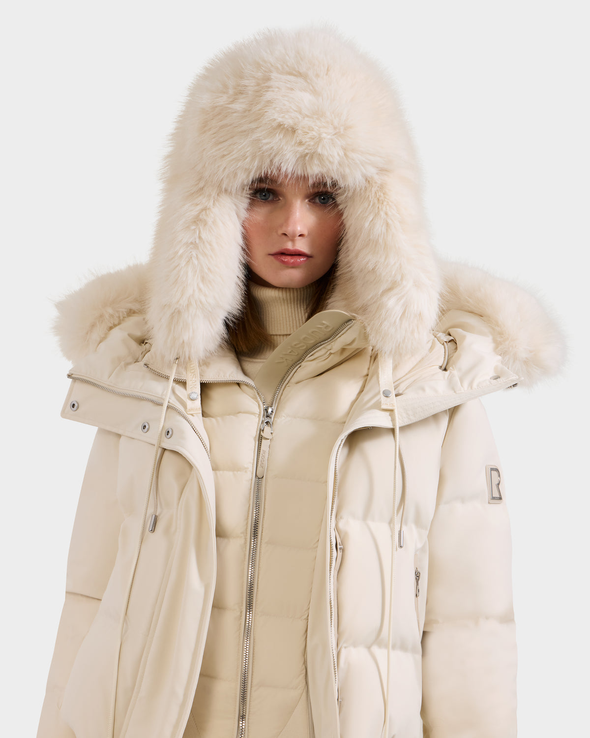 A woman models the Rudsak LIRA - 8425024 Vanilla light beige puffer coat with a fur-trimmed hood and a beige turtleneck, showcasing classic après-ski style as she looks at the camera against a plain background.
