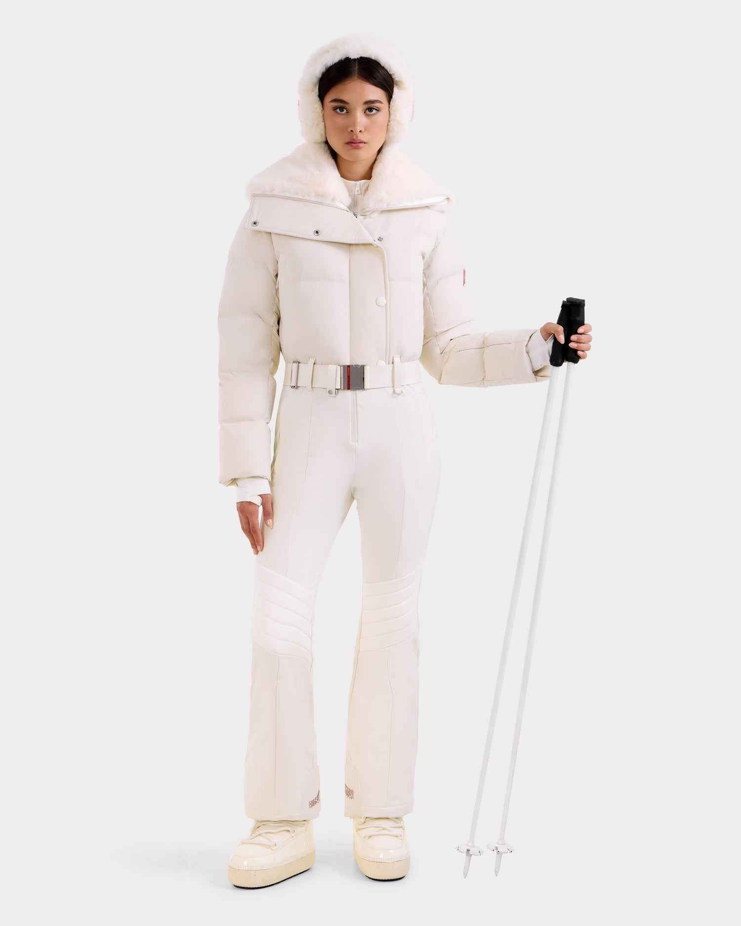 A woman in the Rudsak LOVE - 8124724 Vanilla women's ski suit with a fur-trimmed hood, belted snow pants, puffer jacket, and chunky boots stands holding ski poles against a light background.