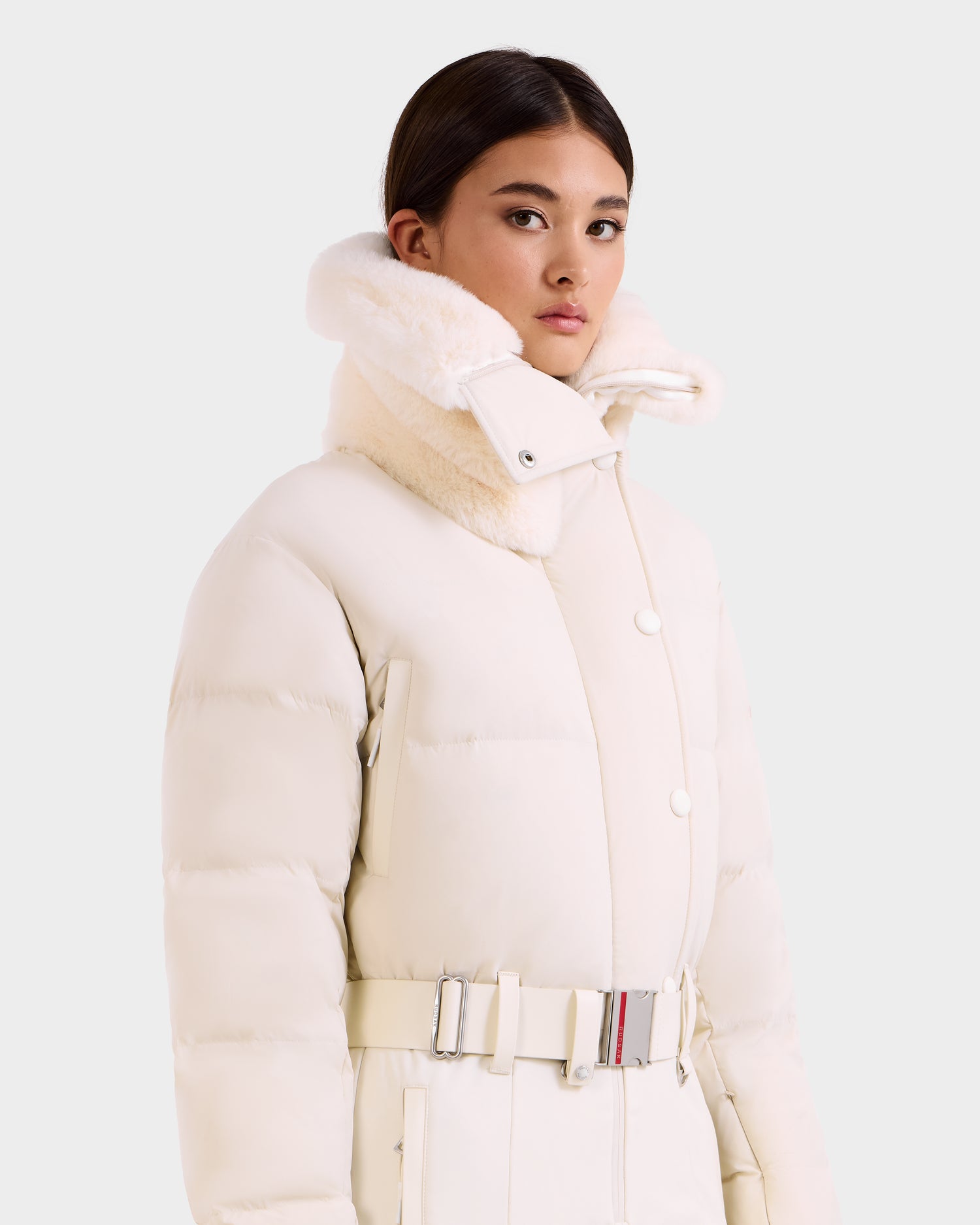 A woman wears the Rudsak LOVE - 8124724 Vanilla puffer coat, featuring an off-white shade, high fur-lined collar, and belted waist, standing against a plain light background.