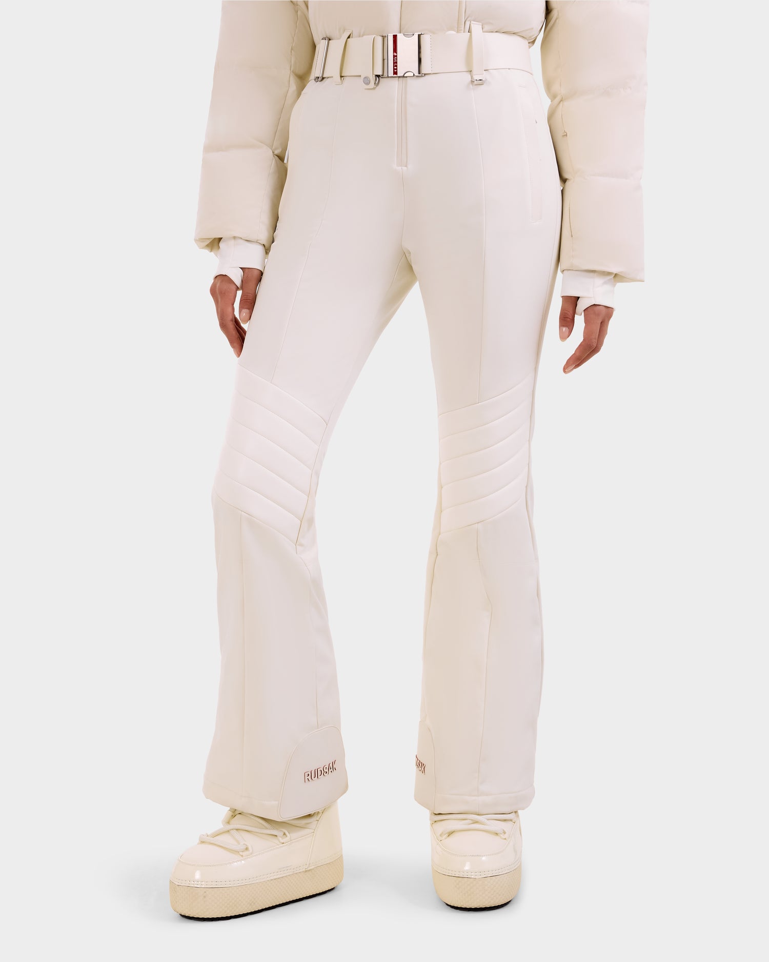 A person wearing the Rudsak LOVE - 8124724 Vanilla cream flared pants with stitched knee details, a matching puffer jacket, belt, and platform boots stands against a plain white background, inspired by the fitted ski suit style.