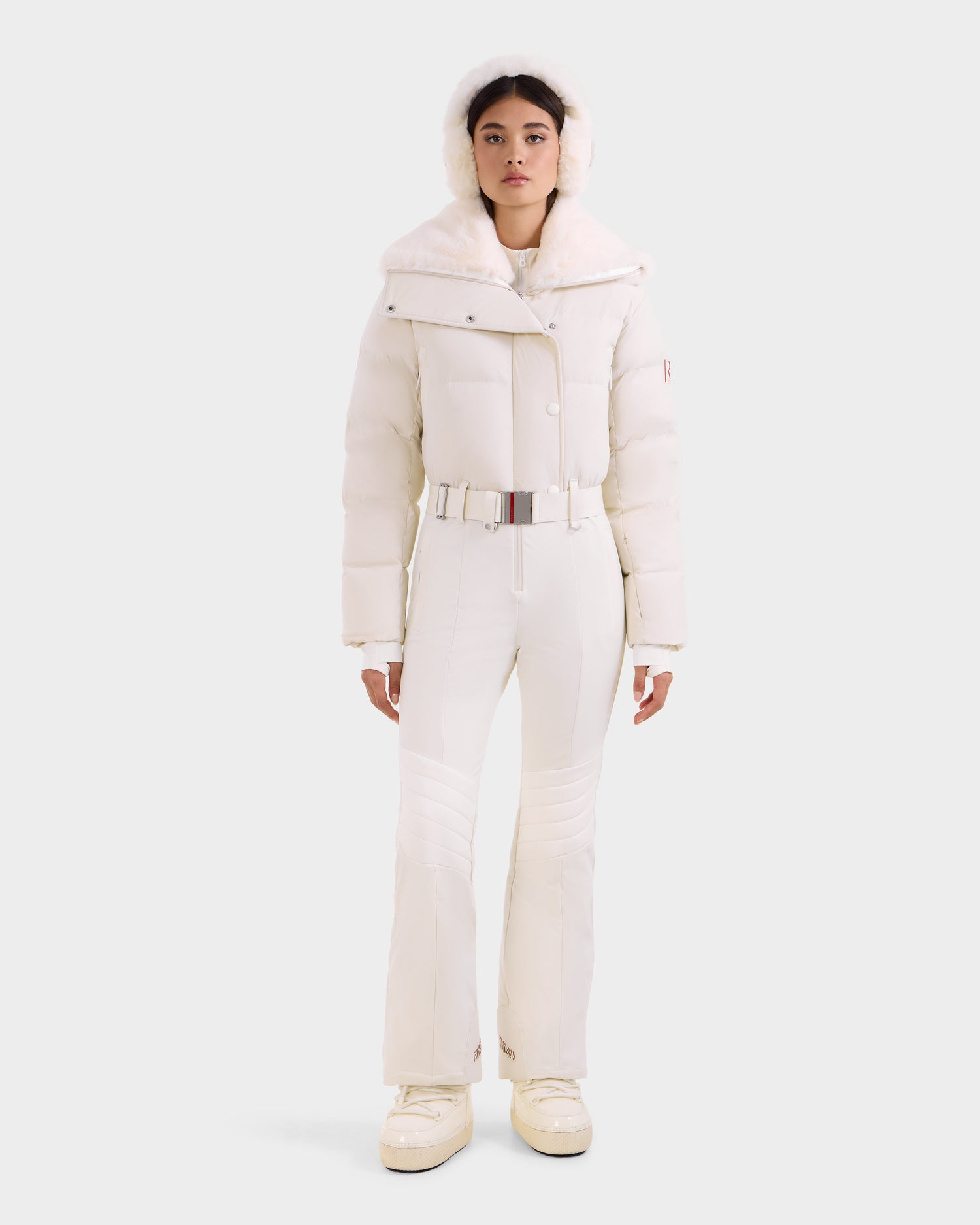 A woman faces forward in the Rudsak LOVE - 8124724 Vanilla women's ski suit, featuring a puffer jacket with RECCO rescue tech, fitted pants, a belted waist, and platform boots, set against a plain light background.