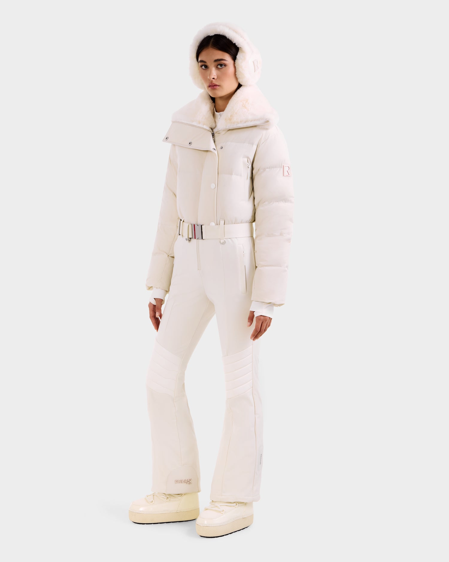 A woman wears the LOVE - 8124724 Vanilla all-white fitted one-piece ski suit by Rudsak, featuring RECCO rescue tech, a fur-lined hood, belt, earmuffs, and platform boots against a light gray background.