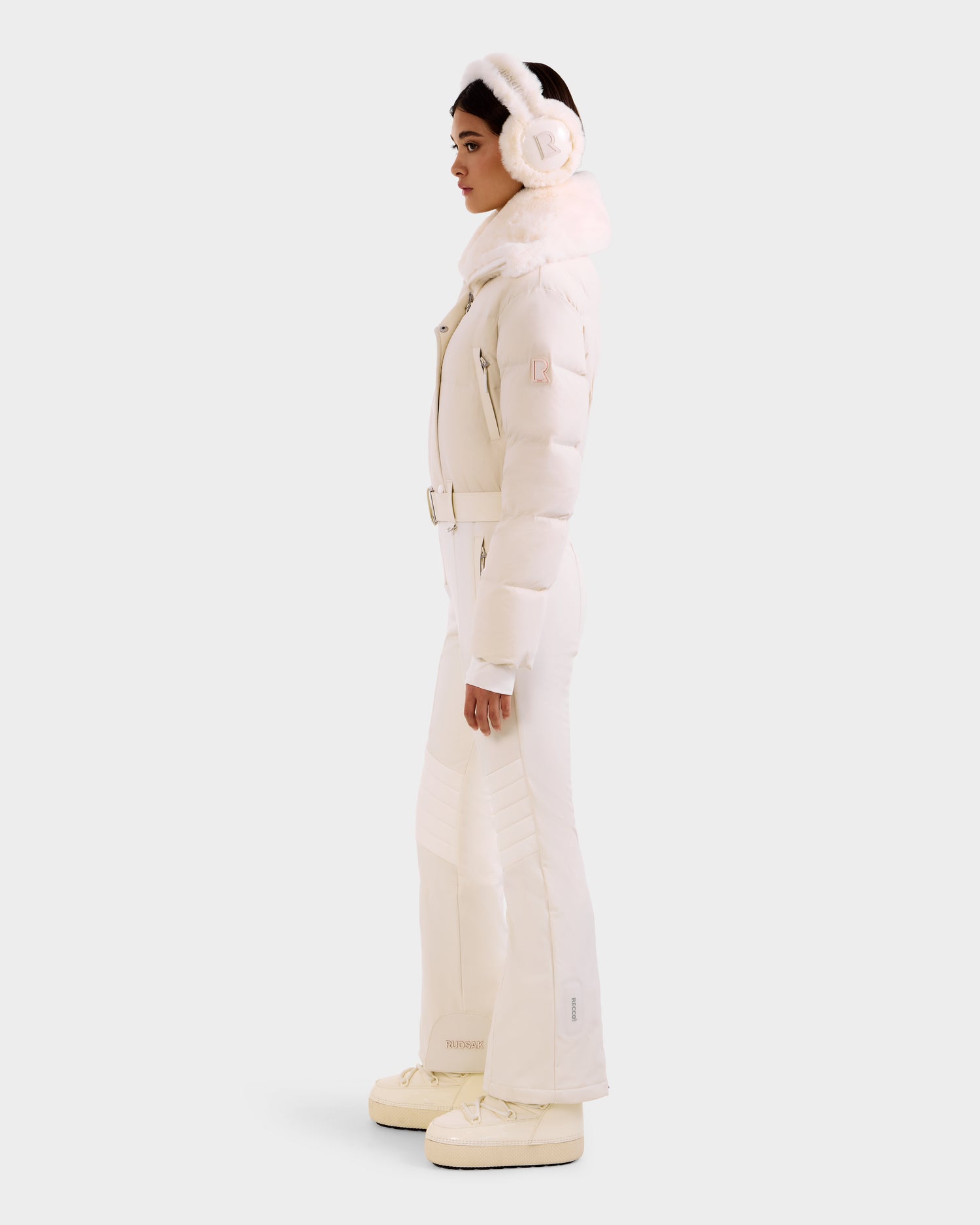 A woman stands in profile wearing the Rudsak LOVE - 8124724 Vanilla women’s ski suit—an all-white set with a puffy jacket, faux fur collar, belted pants, and platform boots—styled with earmuffs against a plain light background.