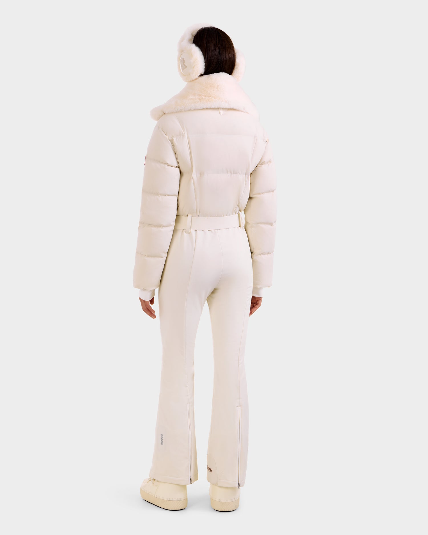 A person faces away wearing Rudsak's LOVE - 8124724 Vanilla women's ski suit with RECCO tech, furry-collared puffer jacket, earmuffs, flared pants, and platform boots, all in white, set against a plain light background.