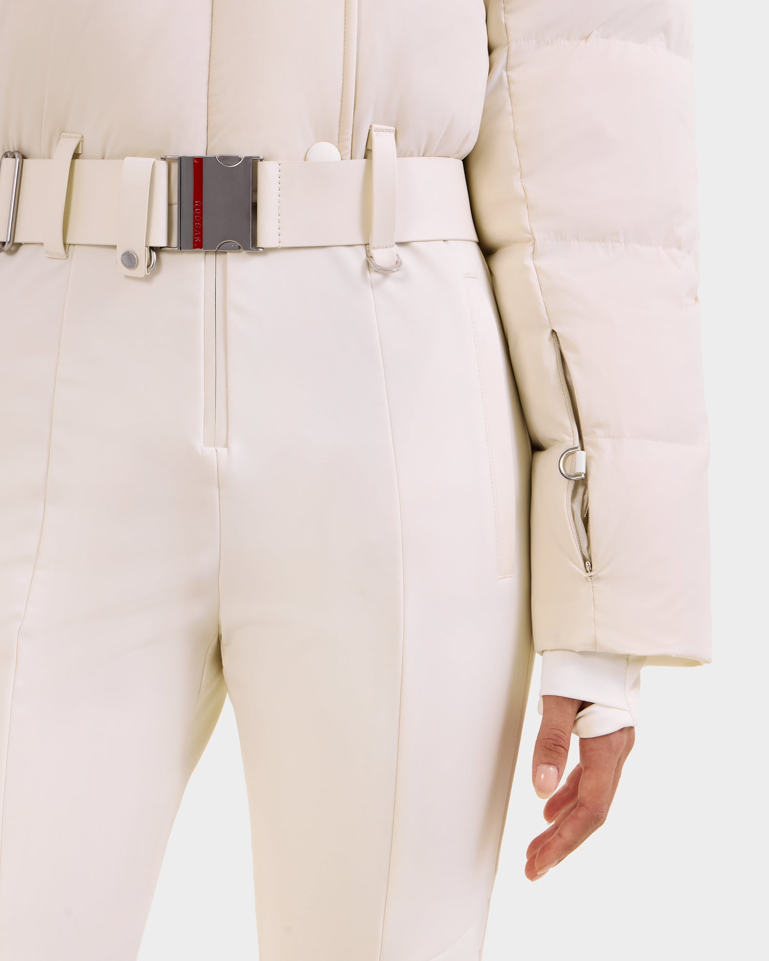 A person is seen wearing the Rudsak LOVE - 8124724 Vanilla women's ski suit, featuring a cream puffer jacket top, matching high-waisted pants, and a wide belt with silver clasp and red-black stripe. Only the torso and right hand are visible.