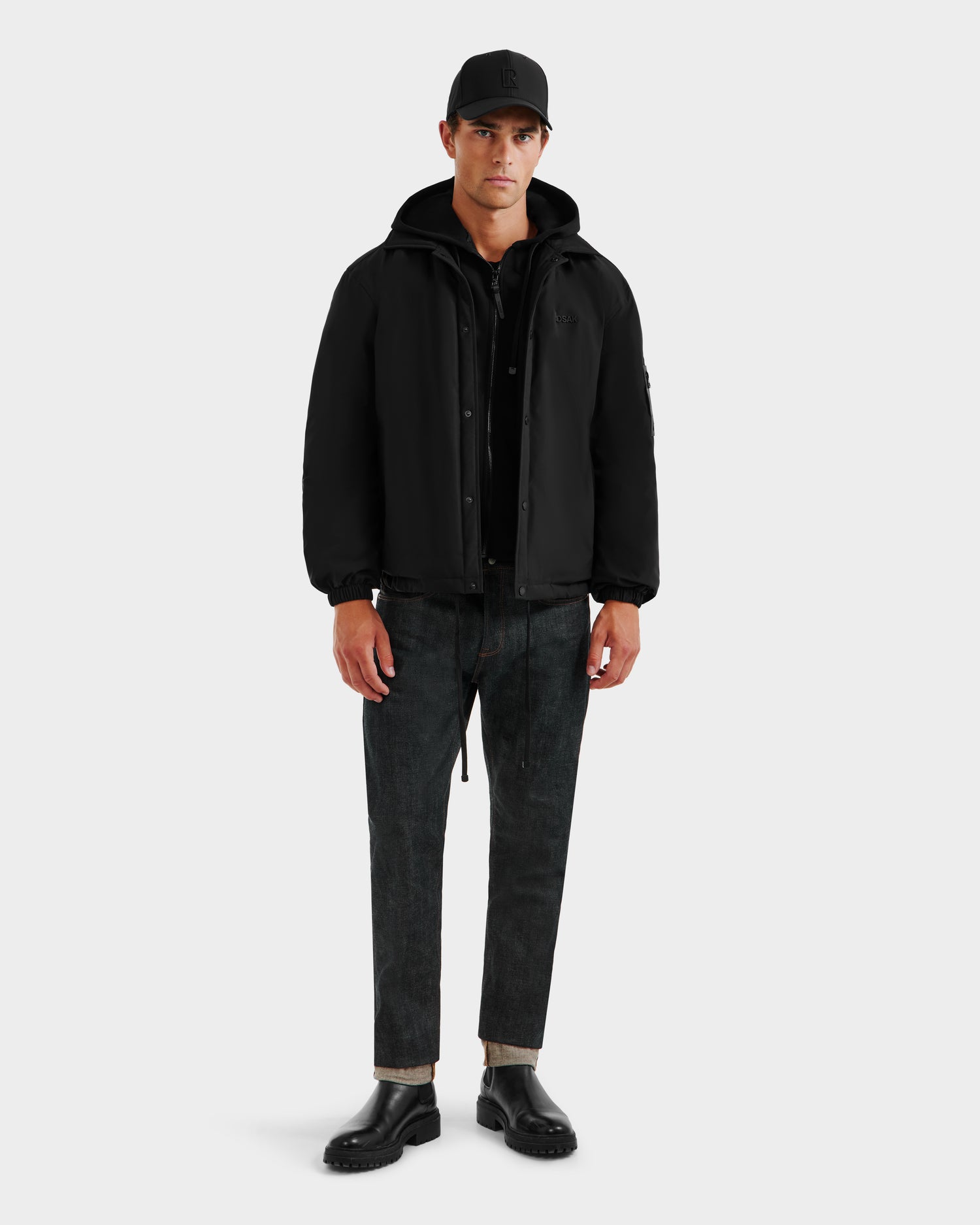 A man stands facing forward, wearing the Rudsak LUCA - 6125009 Black men’s jacket with a lightweight insulated hood, paired with dark jeans and black shoes against a plain light background.