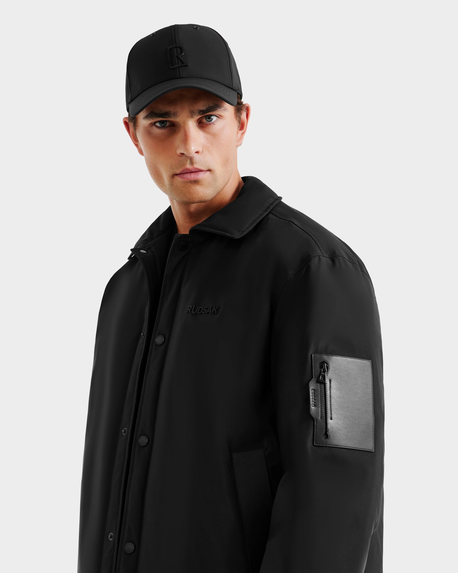 A man wears the Rudsak LUCA - 6125009 Black lightweight insulated men’s jacket with a zippered sleeve pocket and a black baseball cap, standing against a light gray background, looking at the camera with a neutral expression.