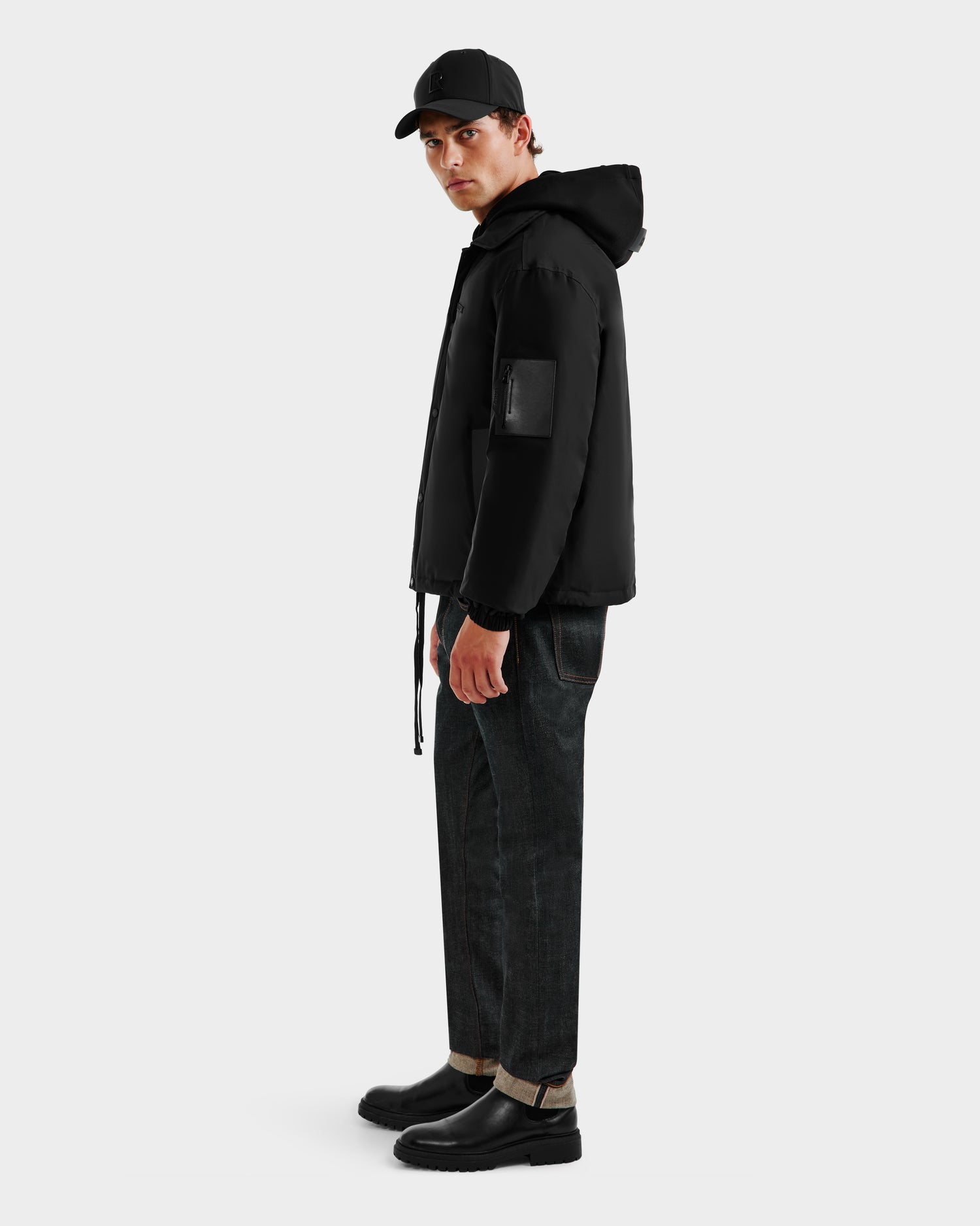 A man in the Rudsak LUCA - 6125009 Black weather-resistant hooded jacket, black cap, dark cuffed jeans, and black boots stands angled to the side against a plain white background.