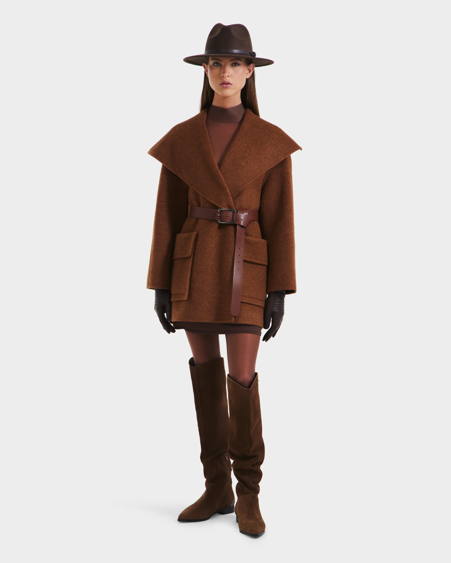 A woman wearing the Rudsak LUCIA - 8125633 Camel jacket with a removable vegan leather belt, matching brown gloves, knee-high boots, a fitted dress, and a brown fedora stands against a plain white background.