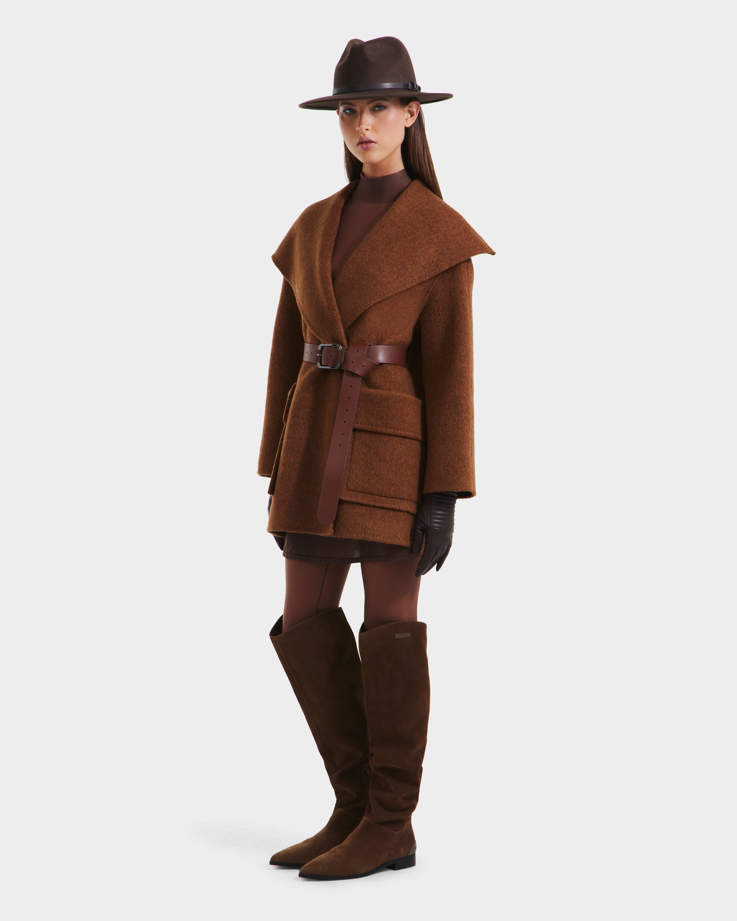 A woman wearing the Rudsak LUCIA - 8125633 Camel womens transitional wool jacket with a removable vegan leather belt stands against a plain white background, styled with brown gloves, boots, a turtleneck dress, and a wide-brimmed hat.
