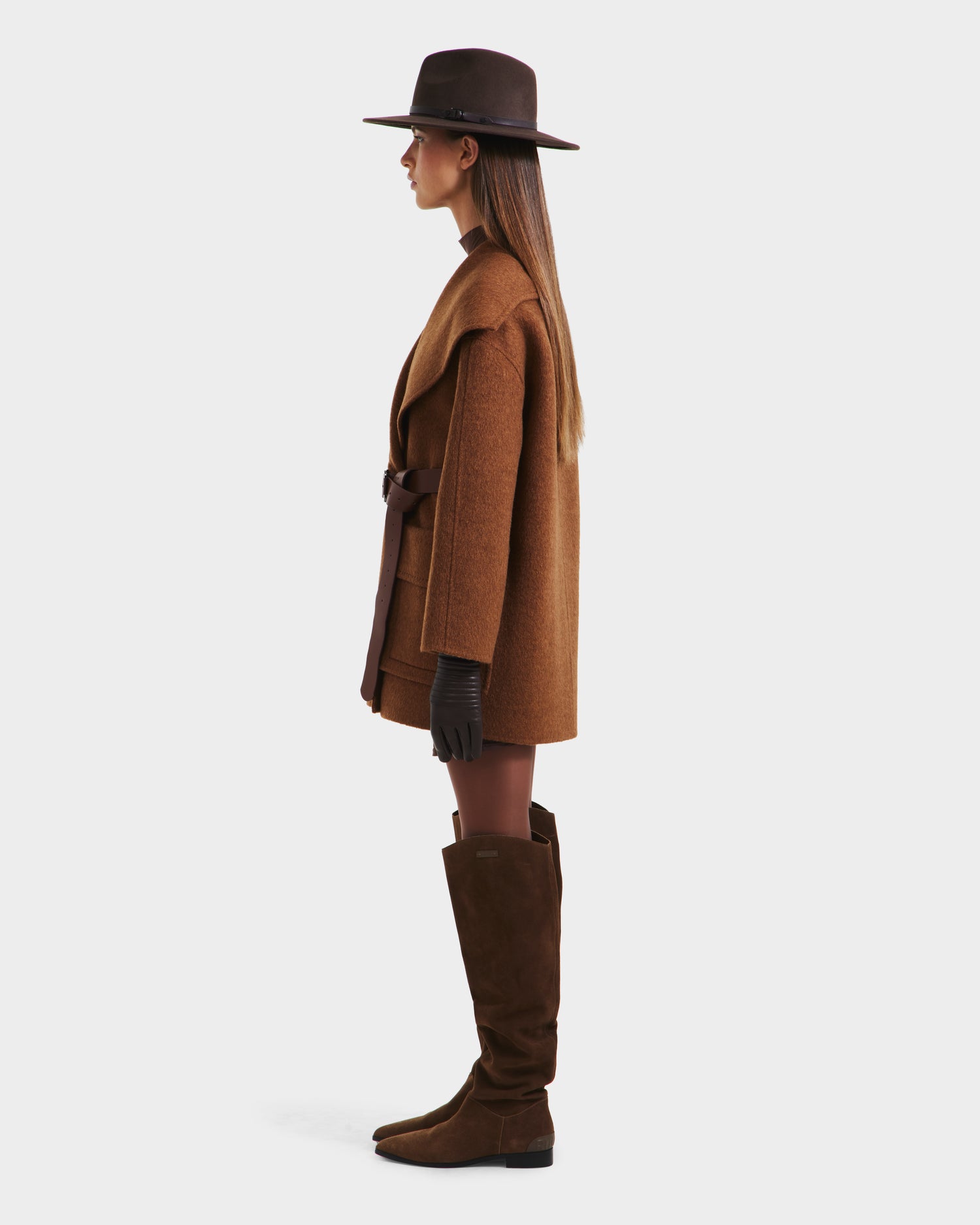 A woman in profile wears the Rudsak LUCIA - 8125633 Camel womens transitional wool jacket with a removable vegan leather belt, wide-brimmed brown hat, dark gloves, and knee-high brown suede boots against a neutral background.