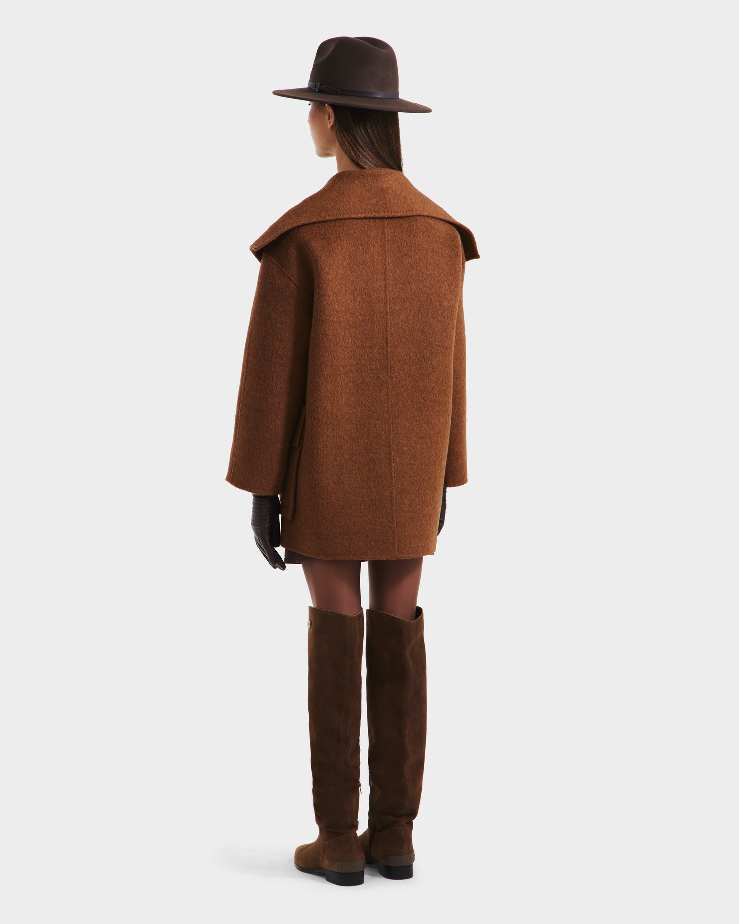 A person in the Rudsak LUCIA - 8125633 Camel womens transitional wool jacket with a removable vegan leather belt, wide-brimmed hat, black gloves, and knee-high brown boots stands facing away against a plain white background.