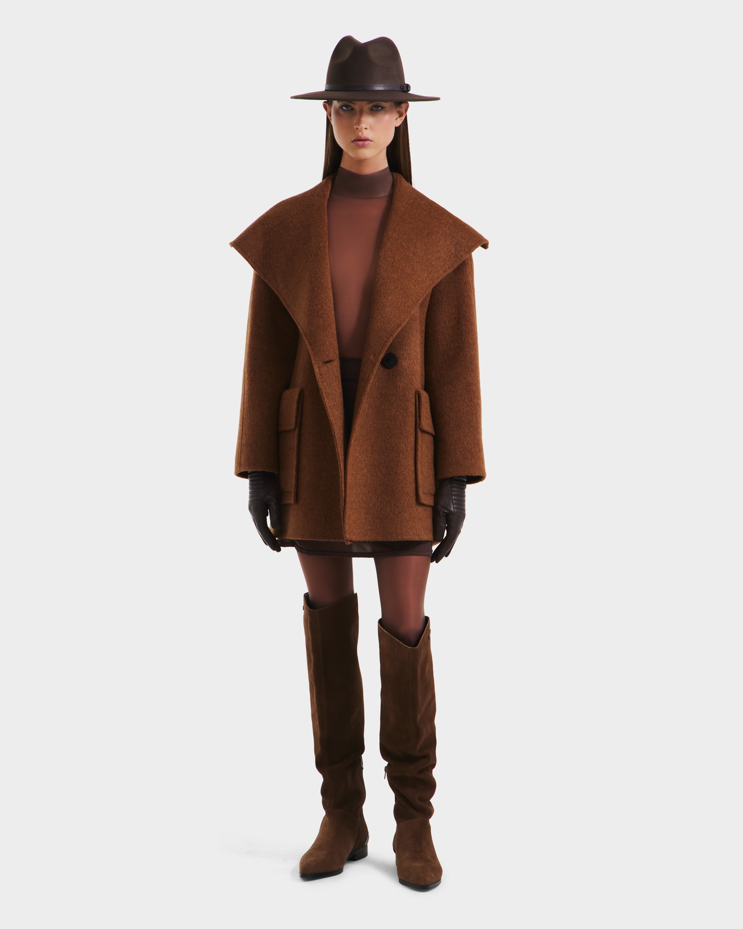 A woman faces forward against a plain white background wearing the Rudsak LUCIA - 8125633 Camel, an oversized womens transitional wool jacket with a removable vegan leather belt, styled with brown accessories and knee-high boots.