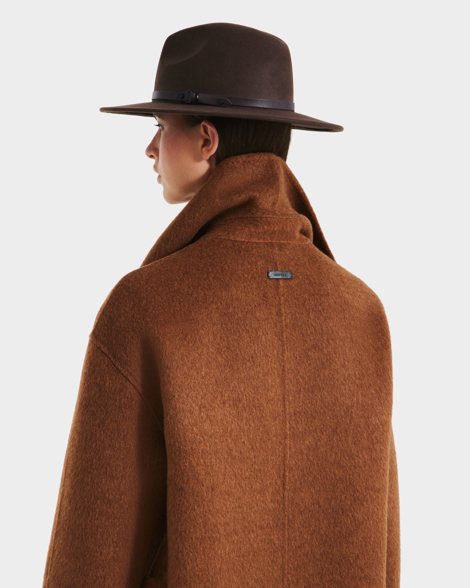 A person is shown from the back and side wearing the Rudsak LUCIA - 8125633 Camel womens transitional wool jacket and a wide-brimmed brown hat against a plain light background.
