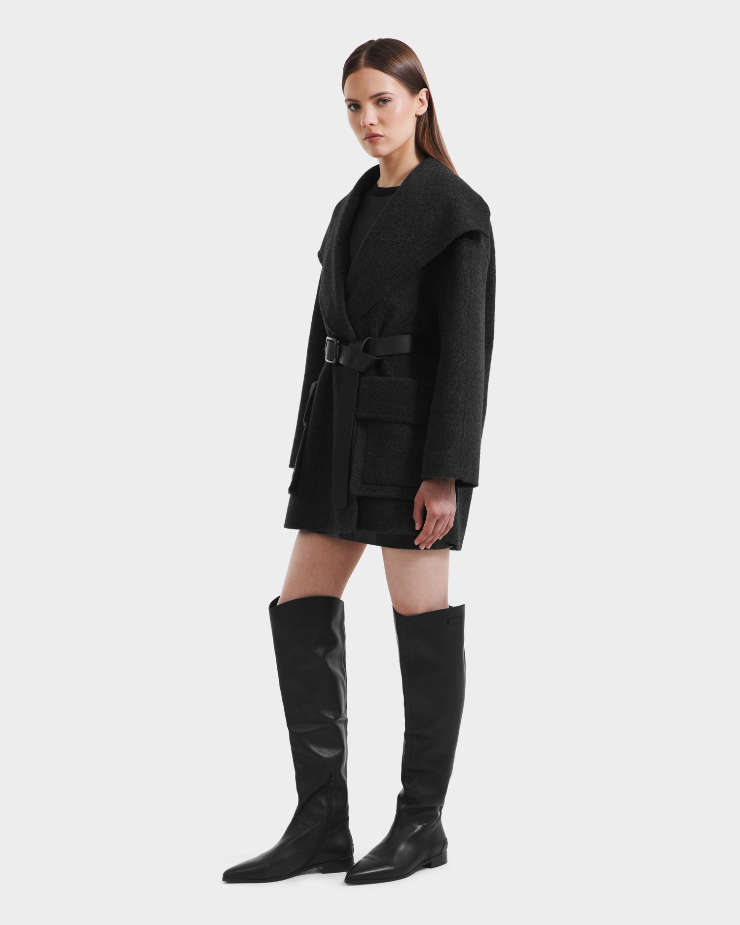 A woman with straight brown hair and a neutral expression models the Rudsak LUCIA - 8125633 Heather Black womens transitional wool jacket with large lapels and a removable vegan leather belt, styled with knee-high black boots against a plain background.