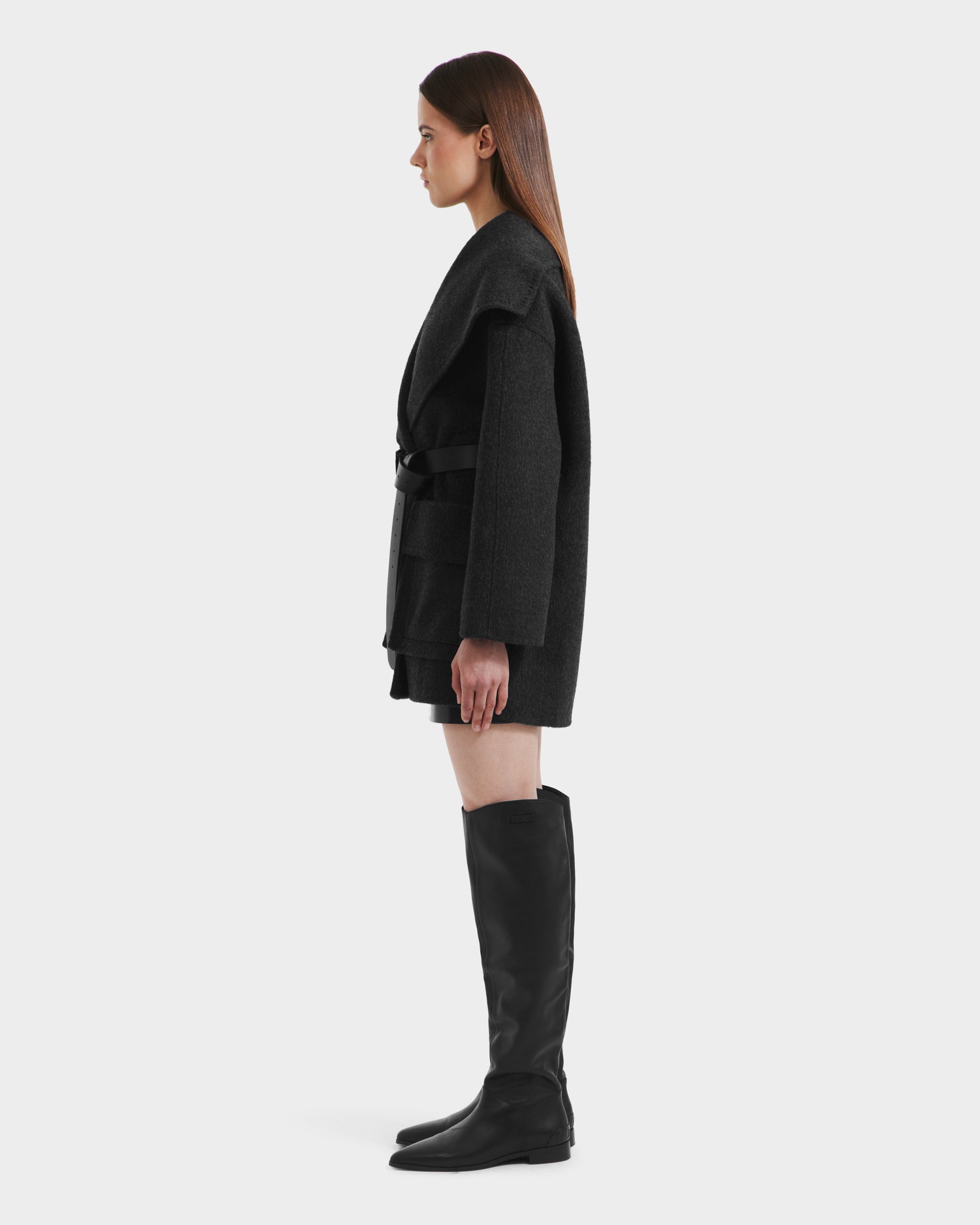 A woman stands in profile wearing the Rudsak LUCIA - 8125633 Heather Black women’s transitional wool jacket with a removable vegan leather belt, paired with black shorts and knee-high black boots against a plain white background.