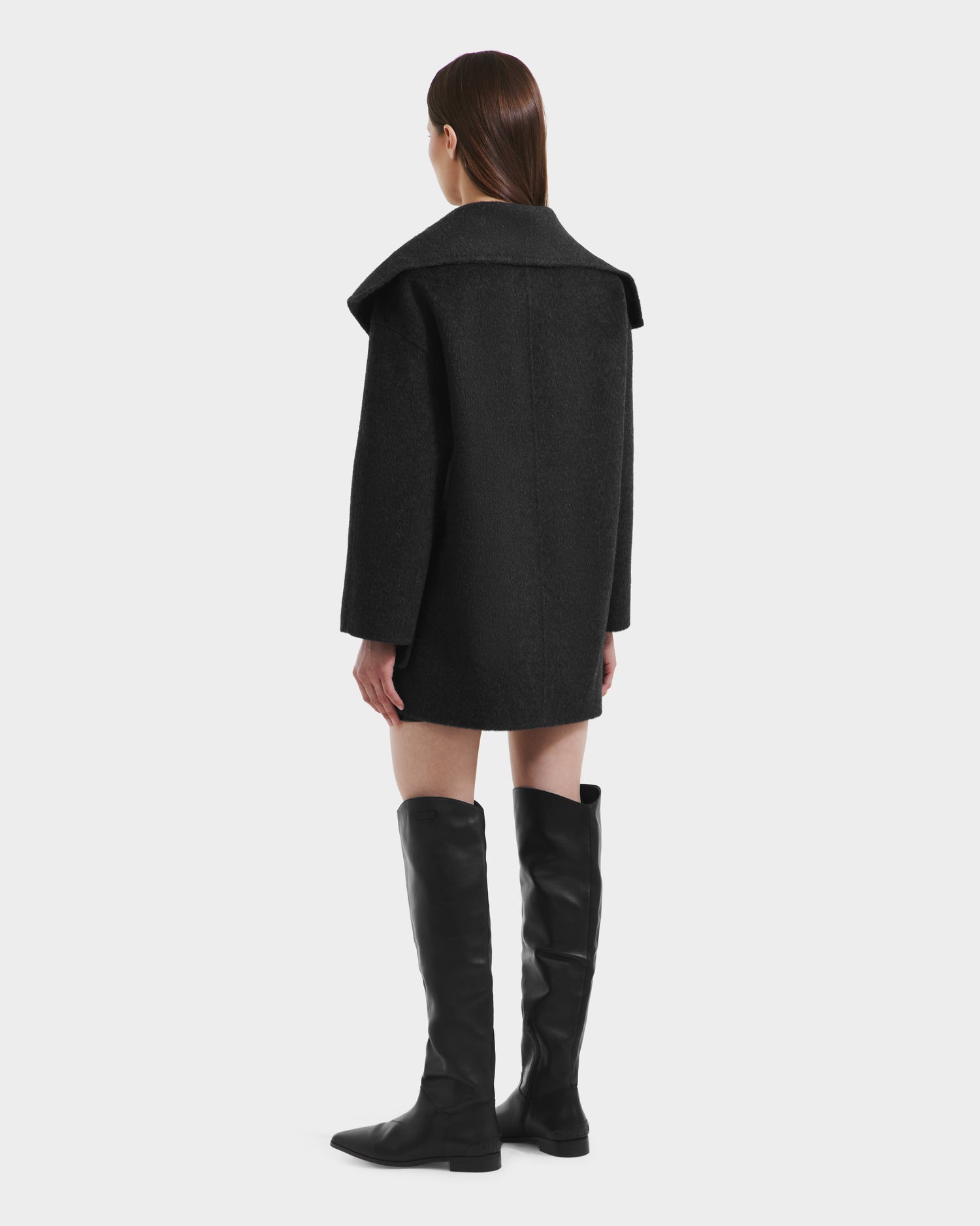 A woman with straight brown hair stands facing away, wearing the Rudsak LUCIA - 8125633 Heather Black transitional wool jacket and knee-high black boots against a plain white background.