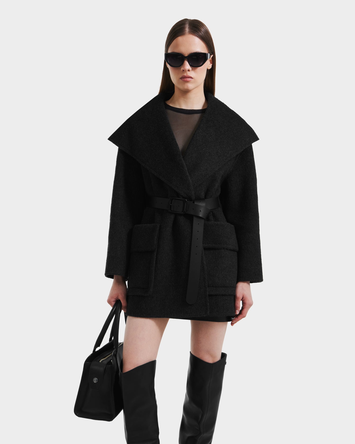 A woman stands against a plain light background wearing the Rudsak LUCIA - 8125633 Heather Black oversized women’s wool jacket with a wide collar, belted waist, knee-high black boots, sunglasses, and holding a black handbag.