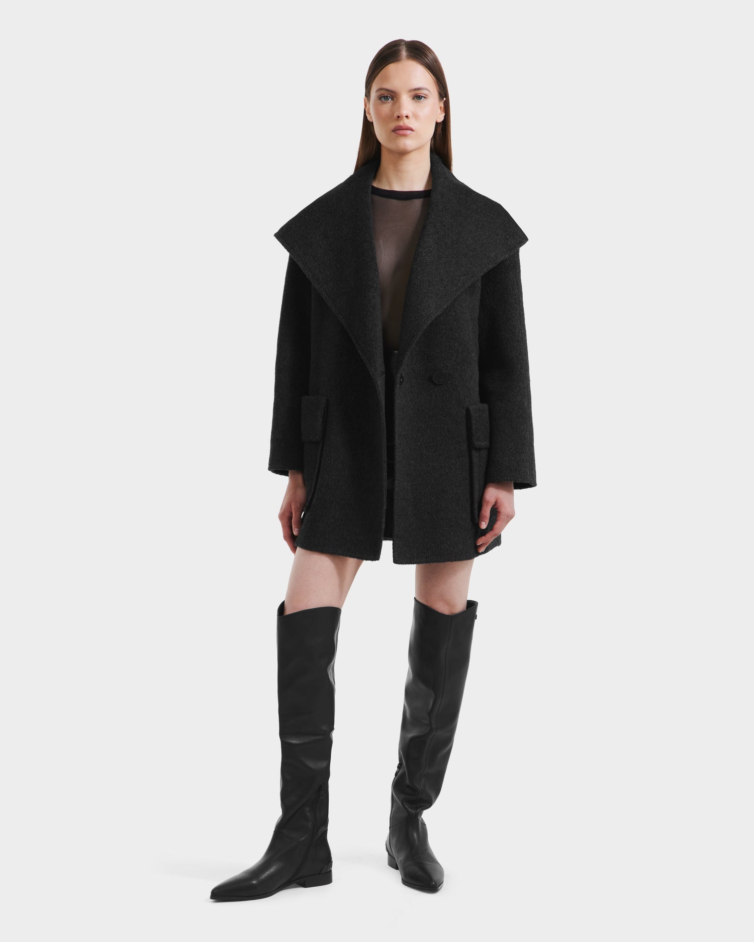 A woman with straight brown hair and a neutral expression stands against a plain background wearing the Rudsak LUCIA - 8125633 Heather Black oversized women’s transitional wool jacket, paired with a sheer black top and knee-high black boots.