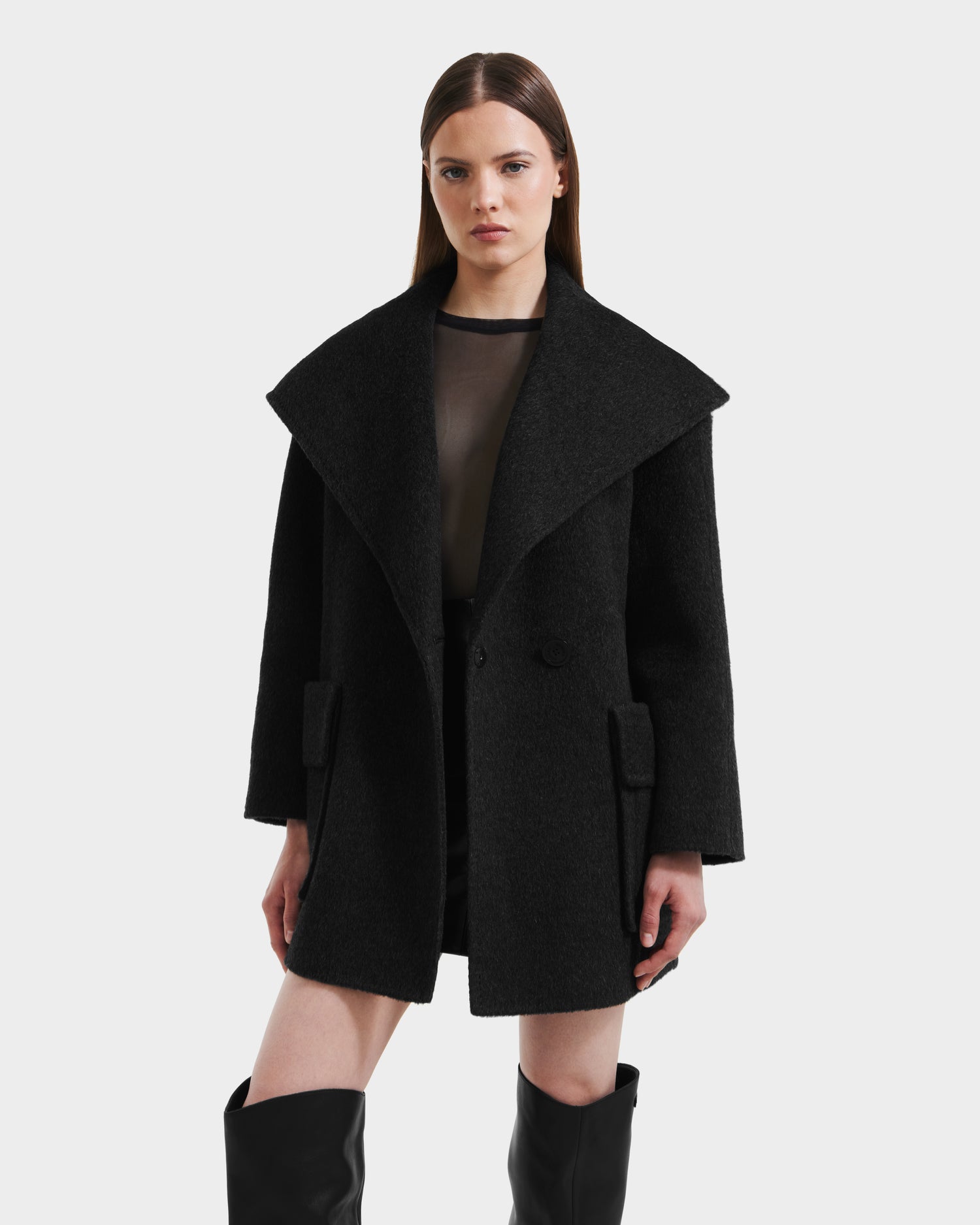 A woman with straight brown hair wears the Rudsak LUCIA - 8125633 Heather Black oversized women’s transitional wool jacket with a wide collar and removable vegan leather belt, styled over a sheer black top and knee-high black boots against a white background.