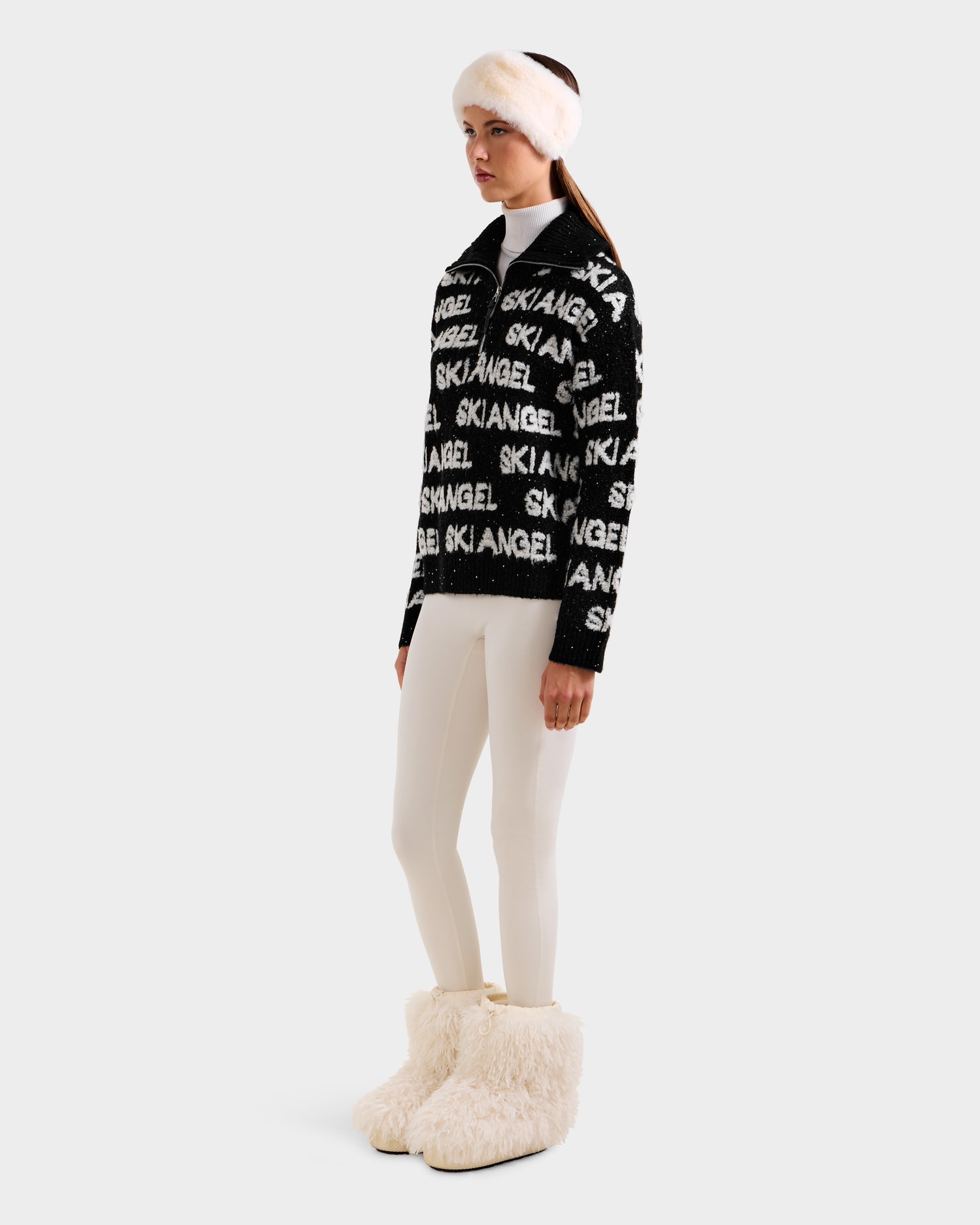 A woman wears the Rudsak LYDIA - 8525687 Black fuzzy après-ski pullover with SKI ANGEL text, styled with white leggings, large white fur boots, and a matching headband against a plain light background.