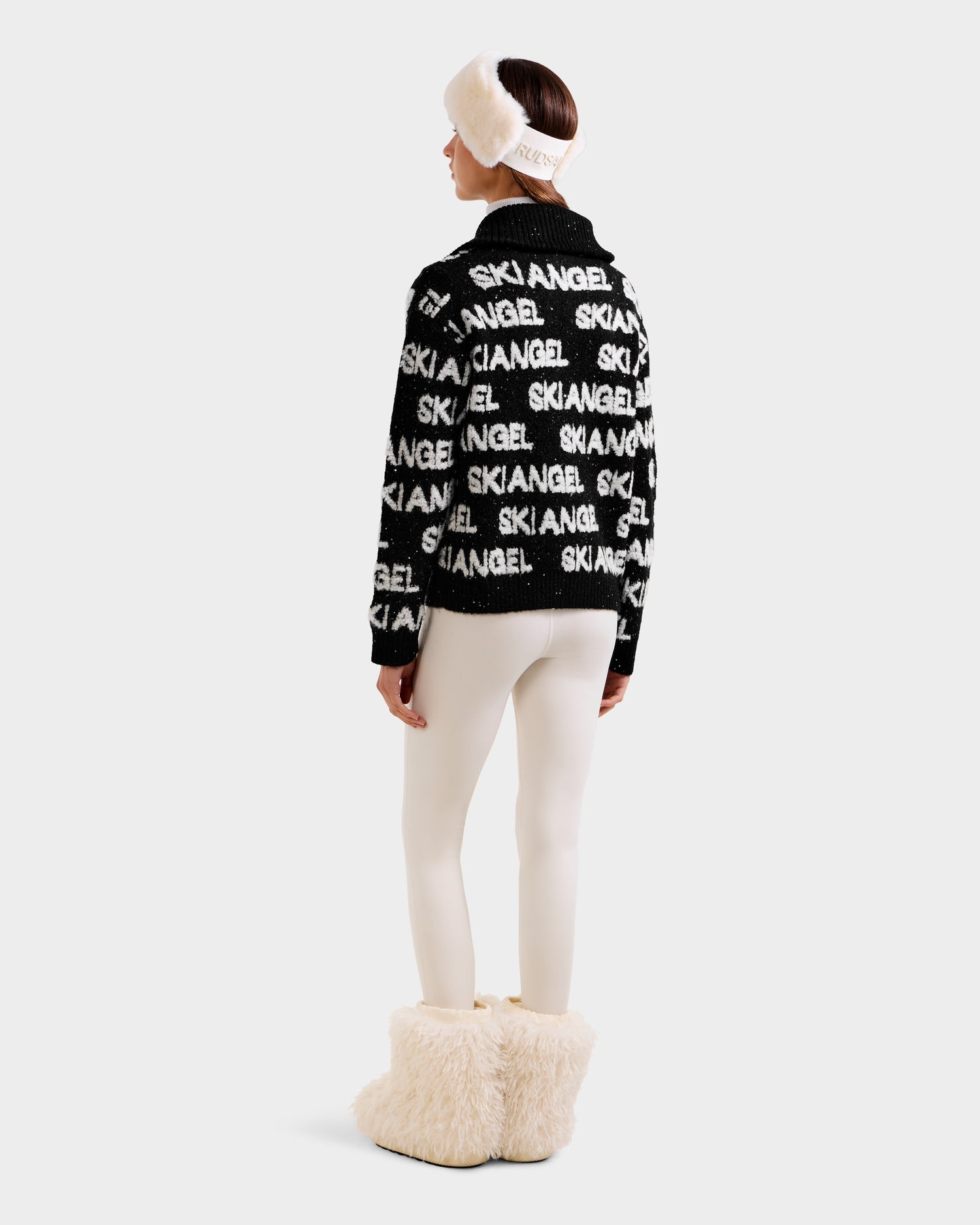 A woman faces away, wearing the Rudsak LYDIA - 8525687 Black jacket with white leggings, cream fluffy boots, and a matching ANGEL headband—her stylish après-ski look stands out against a simple light background.