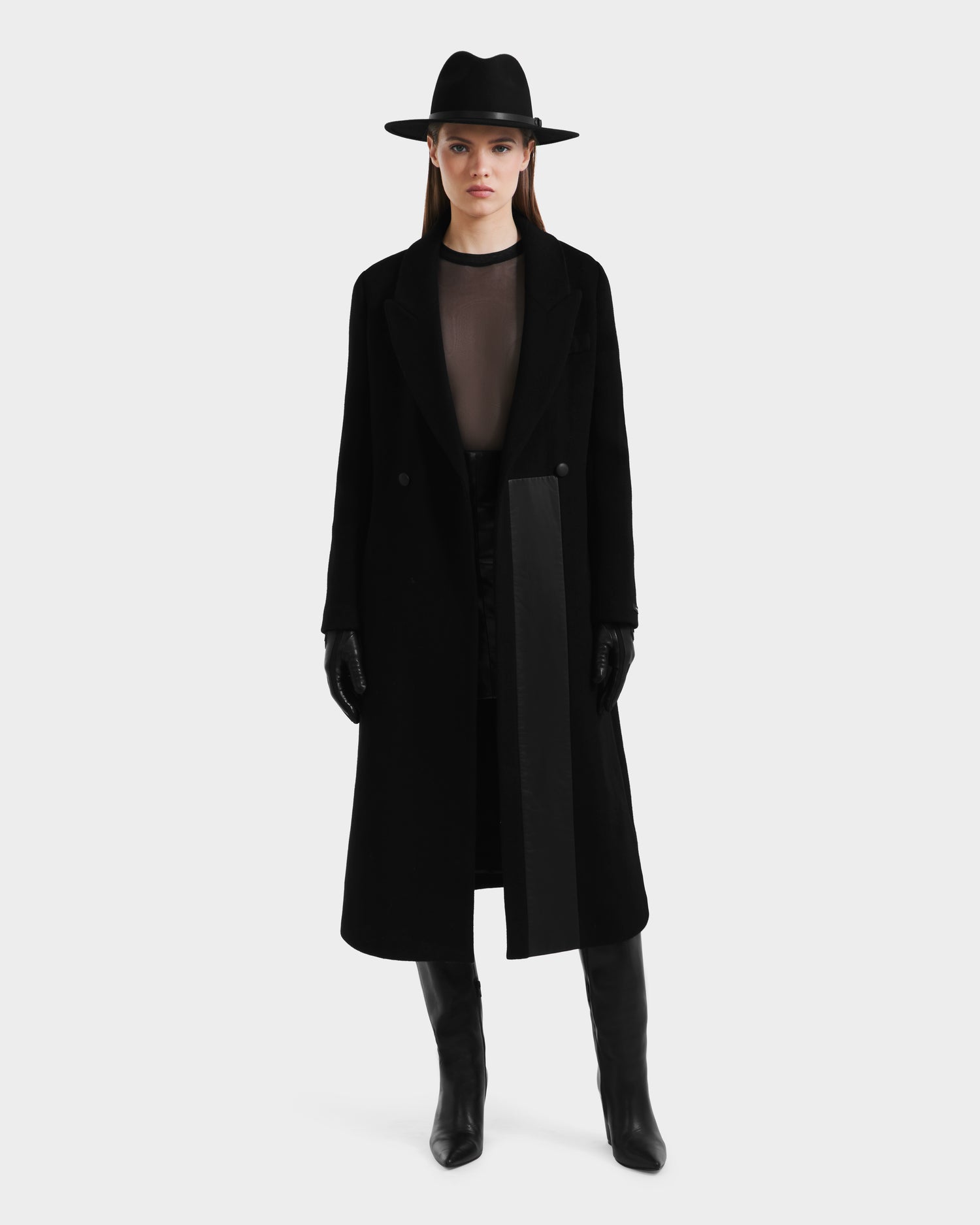 A woman faces forward wearing the Rudsak LYNWOOD - 8125539 Black long tailored coat, paired with black gloves, knee-high boots, a sheer top, and a wide-brimmed hat against a plain light background.