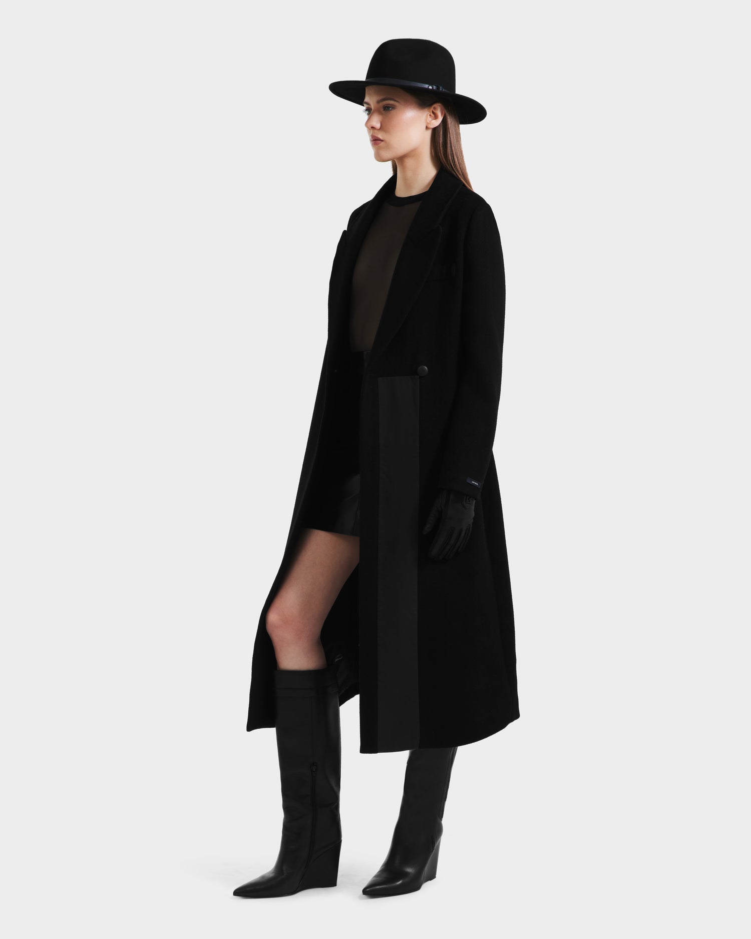 A woman models the Rudsak LYNWOOD - 8125539 Black long tailored coat with a leather front panel, styled with a sheer black top, black shorts, gloves, and knee-high boots against a plain white background.
